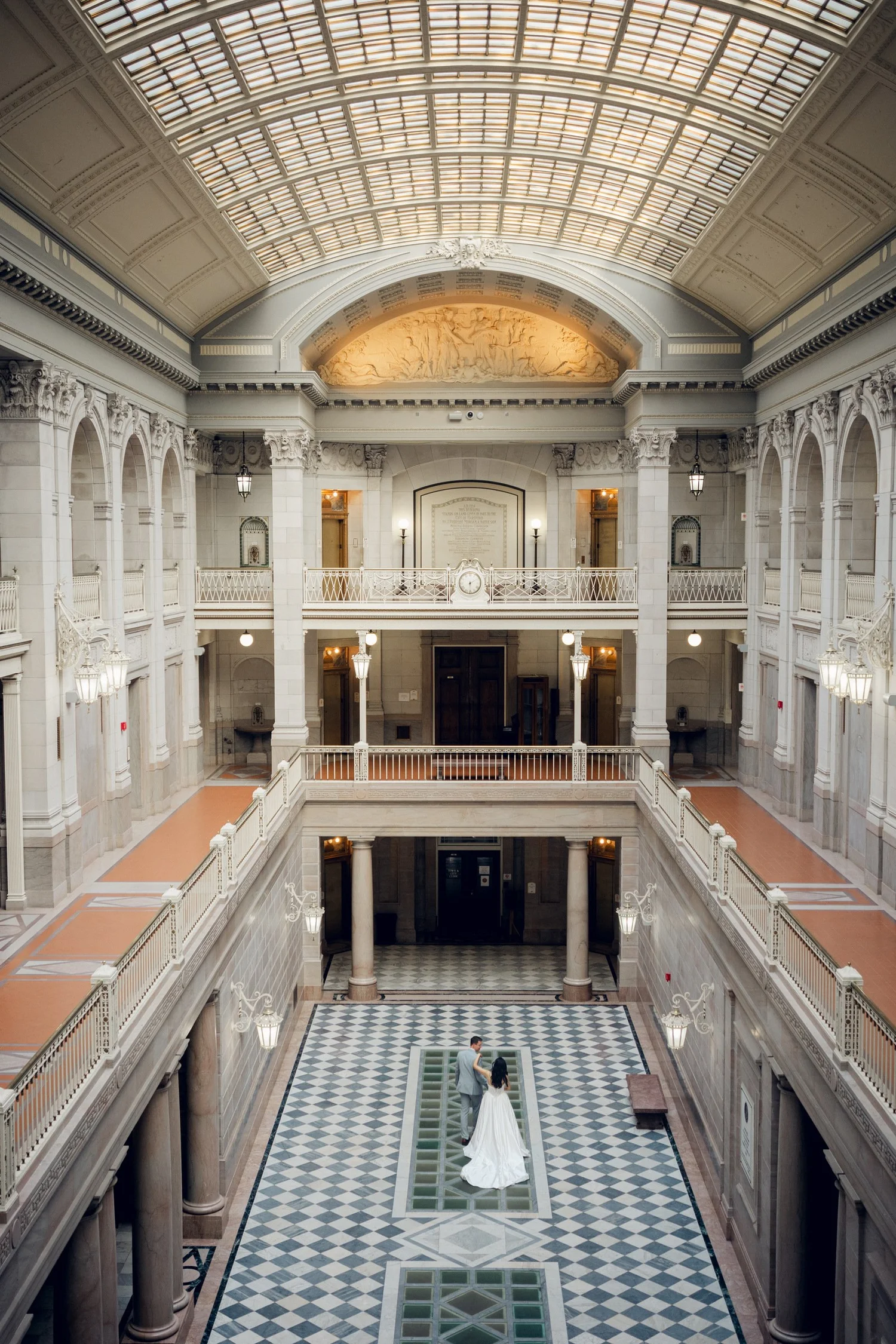Wide architectural wedding portrait showing all three tiers of marble balconies, arched colonnades, and the vaulted skylight inside Hartford City Hall, Hartford, CT
