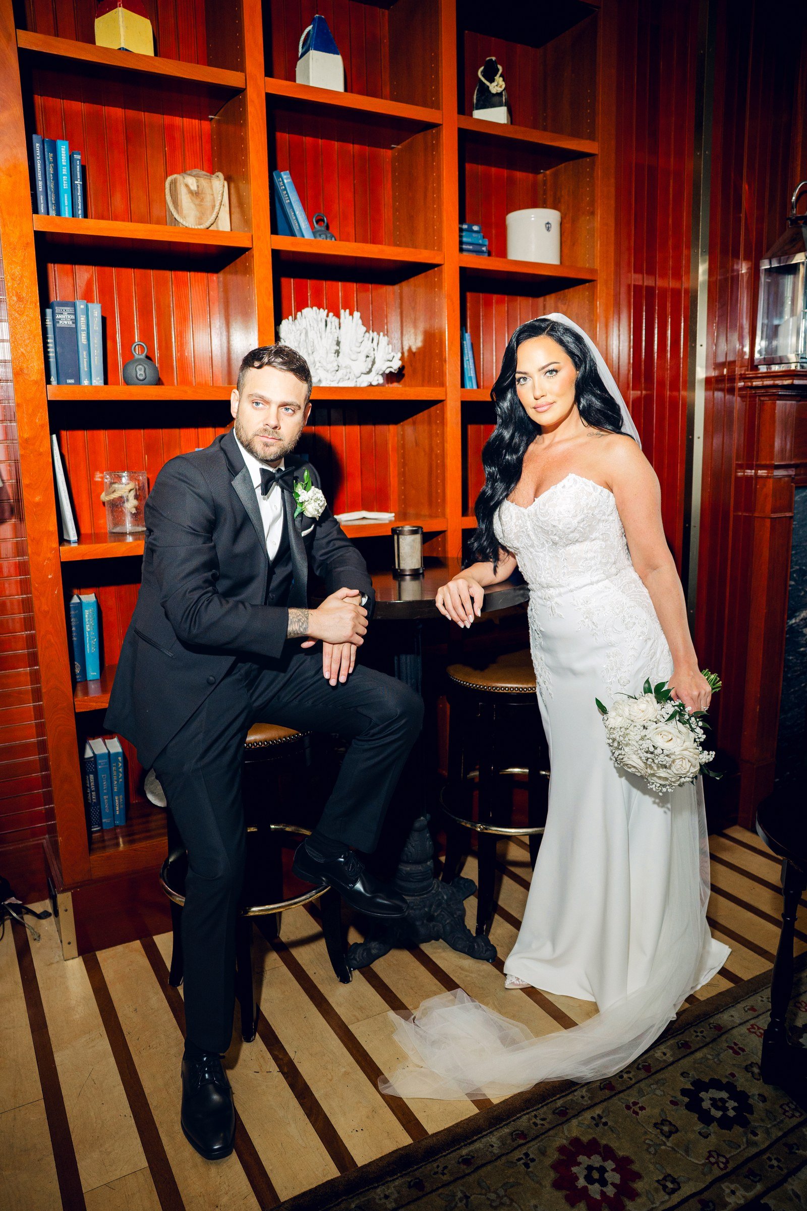 Beauport Hotel wedding videographer, Gloucester, MA — groom seated beside the bride against red library bookshelves