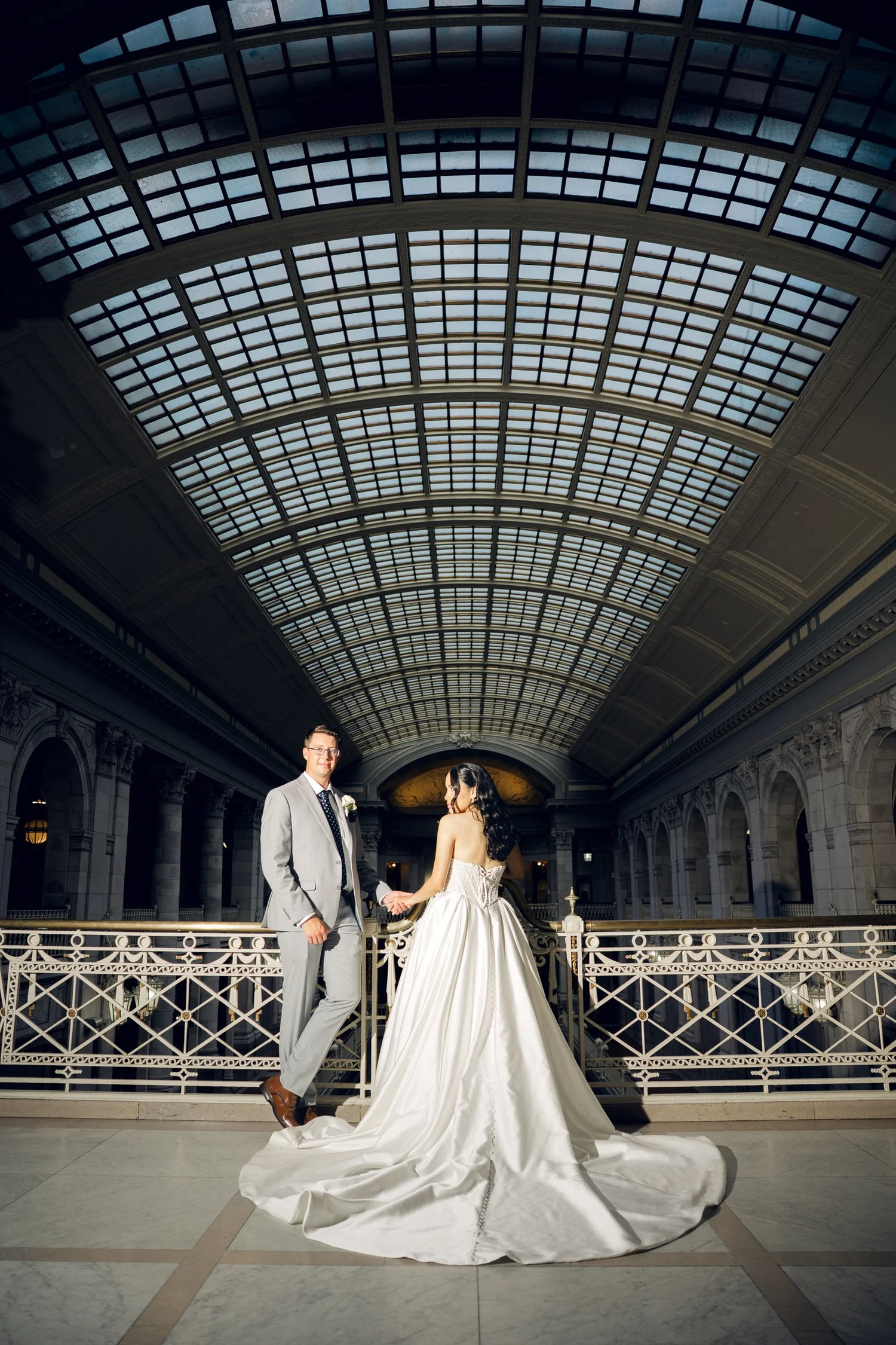 Low-angle wedding portrait under the barrel-vaulted glass skylight ceiling at Hartford City Hall with the bride's cathedral train spread across the marble floor