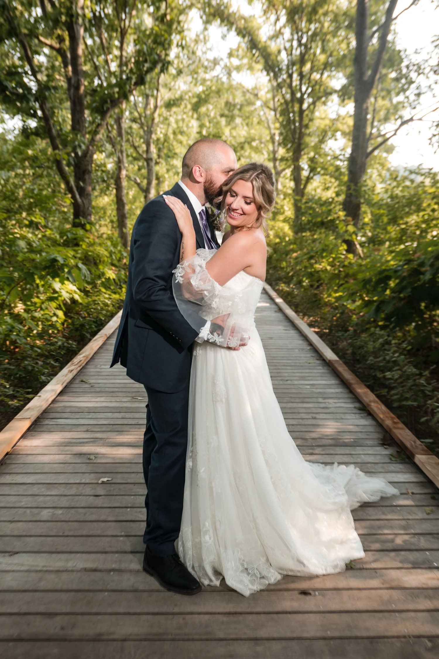 Romantic portrait of the groom kissing the bride's forehead on the wooden boardwalk bridge surrounded by sunlit woodland foliage at Tower Hill Botanic Garden, Boylston, MA