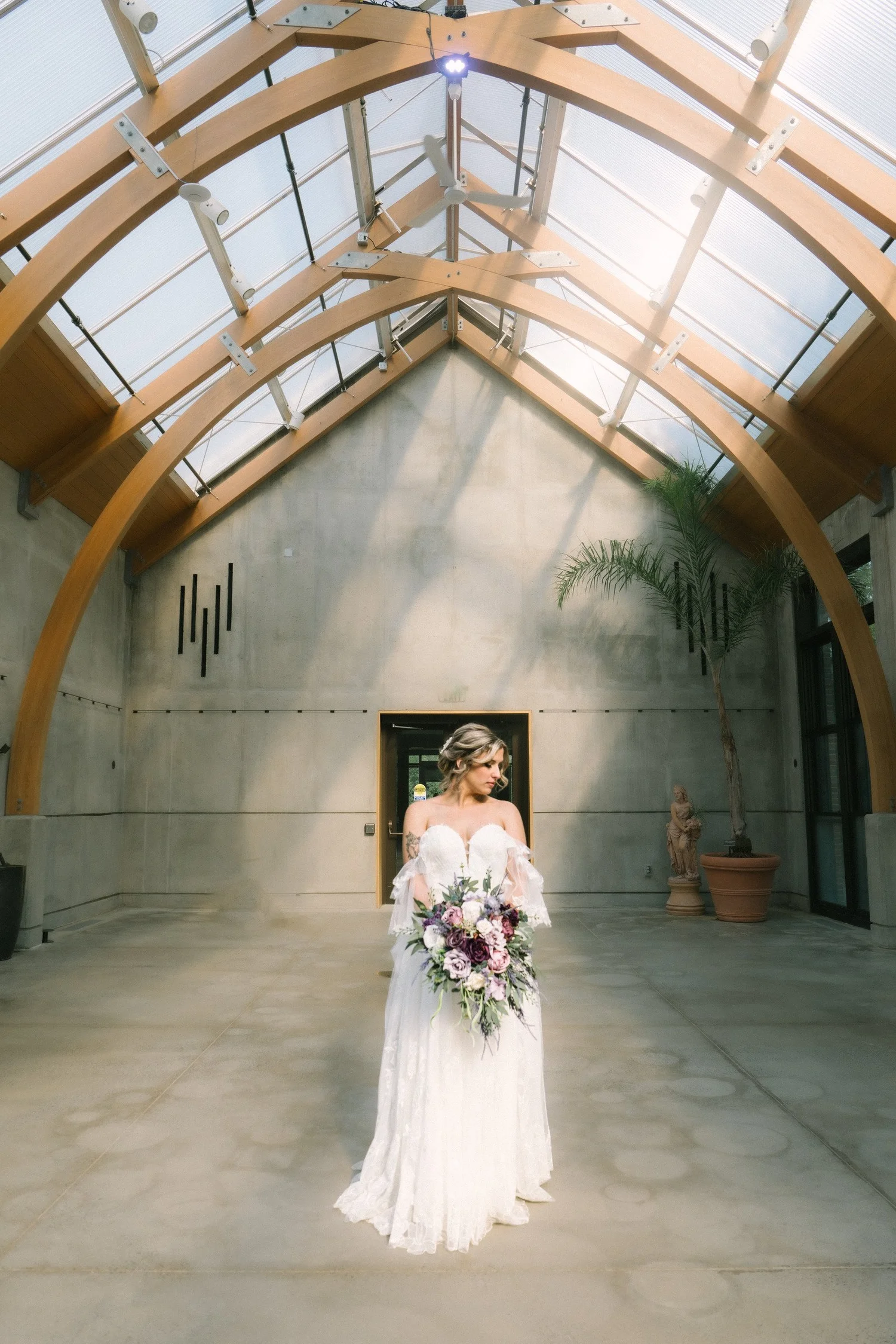 Bride in her wedding gown holding a lavender bouquet beneath the timber-and-glass arches of the conservatory at New England Botanic Garden at Tower Hill, Boylston, MA