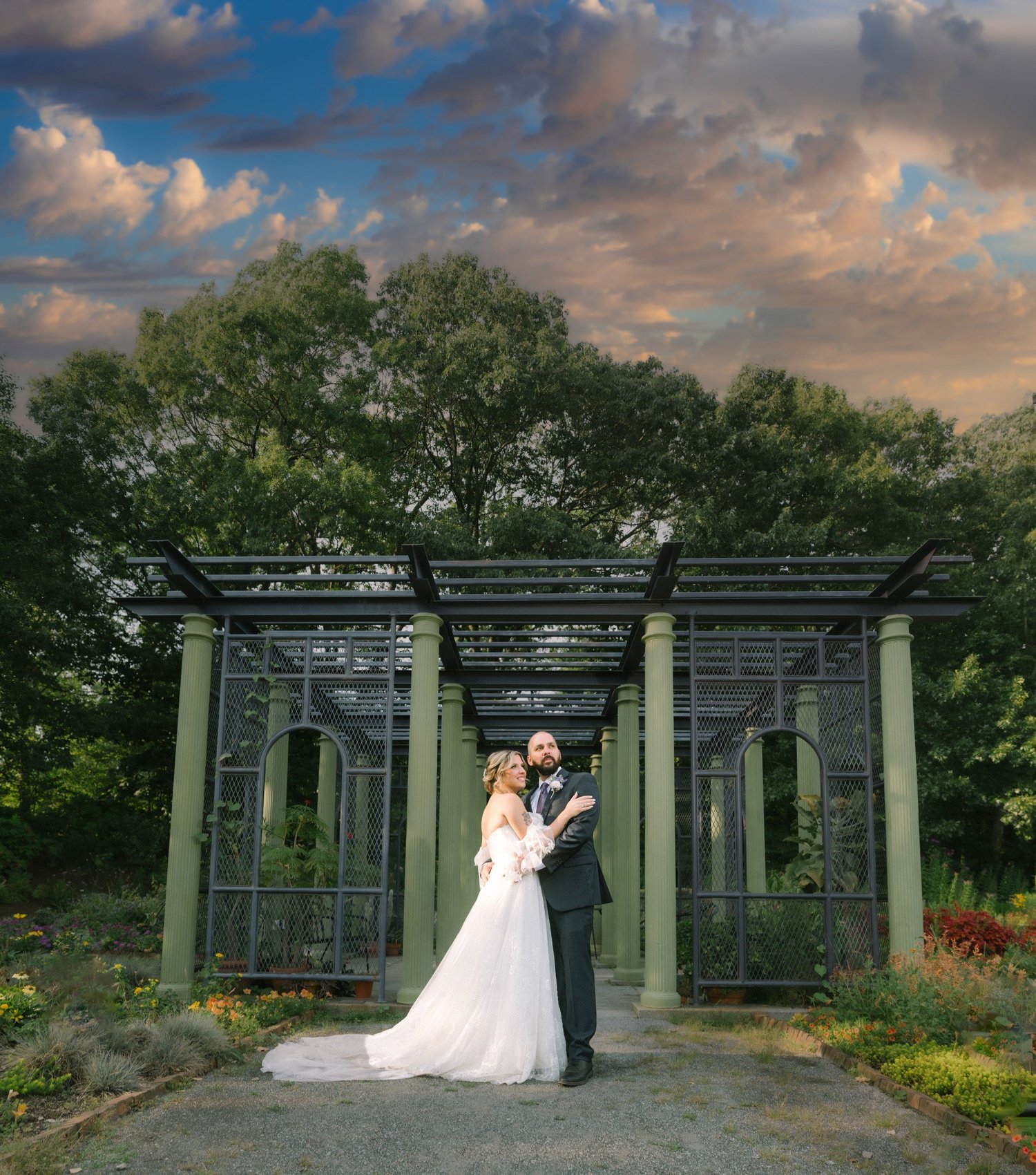 Wide sunset portrait of the bride and groom under the dark green trellis pergola with dramatic clouds at New England Botanic Garden at Tower Hill, Boylston, MA