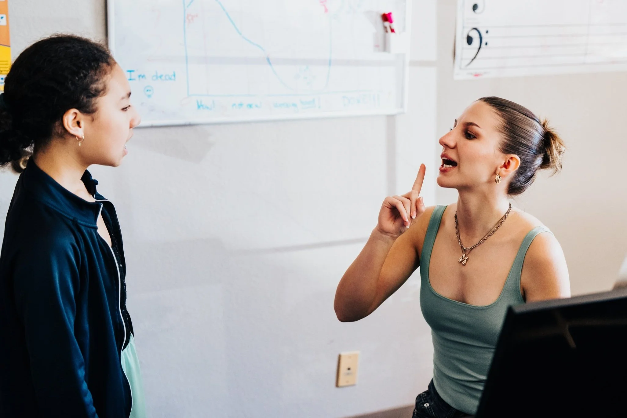 A voice teacher modeling a mouth and facial technique while her student watches and sings
