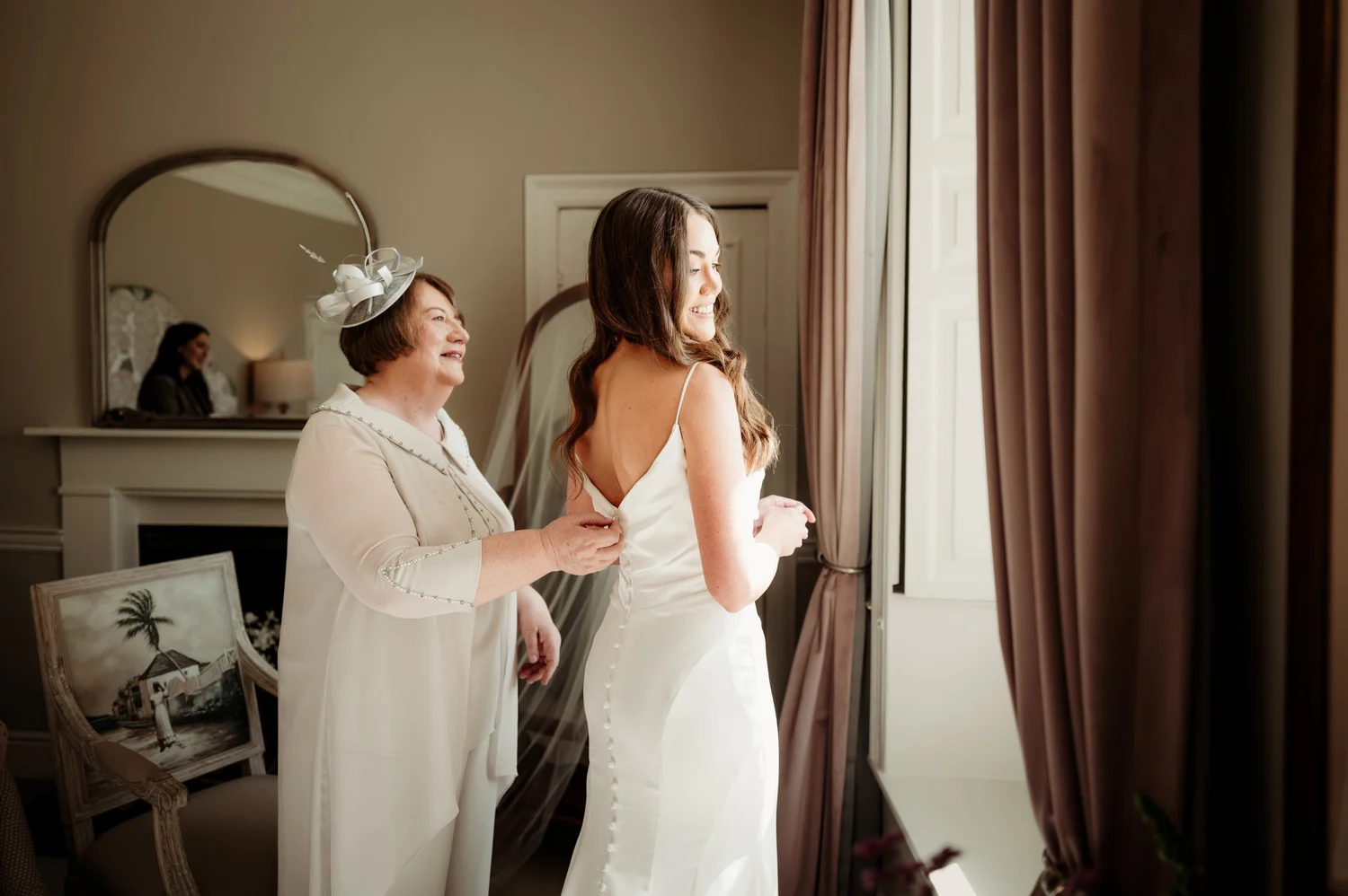 A bride smiling by the window while her mother helps fasten her wedding dress.