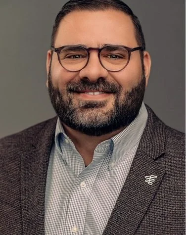 Conference headshot: smiling man with glasses and a beard in a brown blazer on a grey backdrop