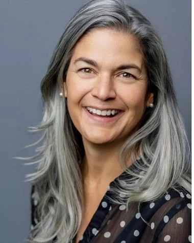 Conference headshot: smiling woman with long silver hair in a polka-dot blouse on a grey backdrop