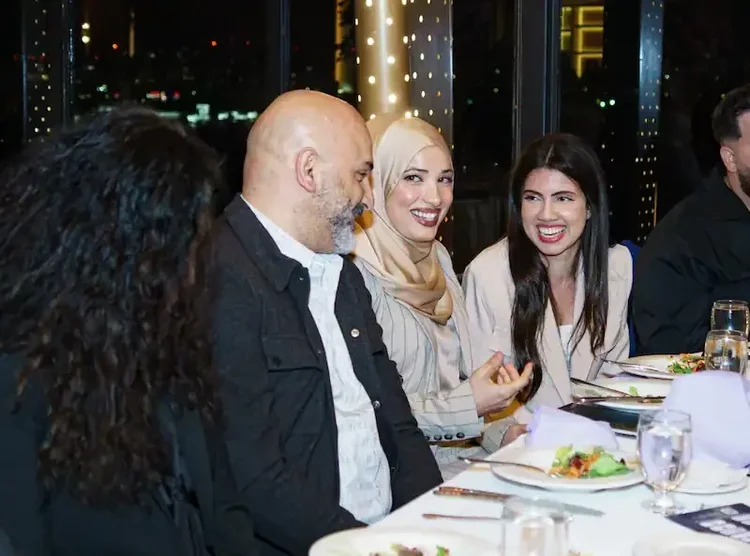 Three guests smiling together at their table, the city skyline lit up through the windows behind them
