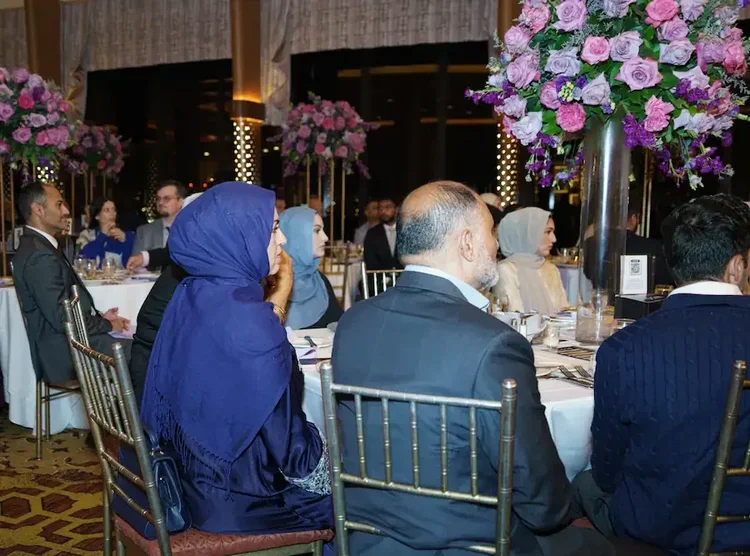 Guests seated at round tables watching the evening's program, floral centerpieces and city windows behind them