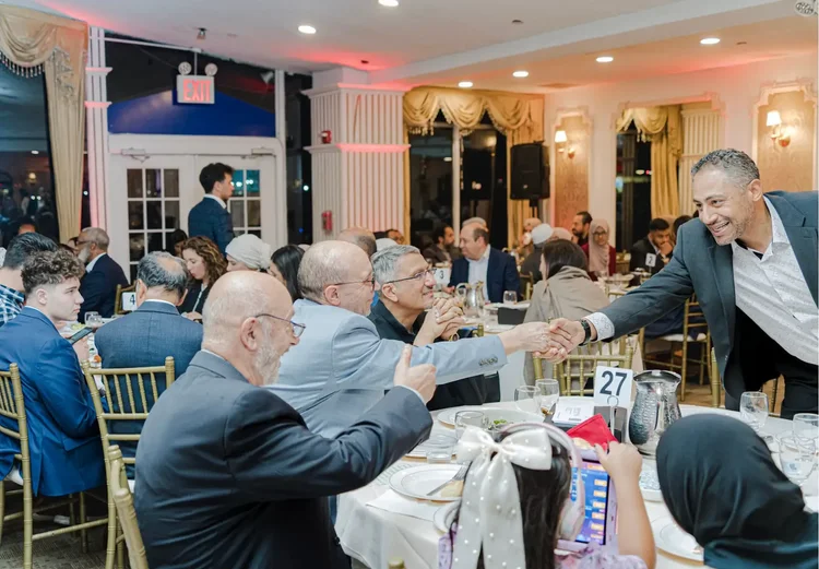 A guest reaching across a dinner table to shake hands with attendees seated around it