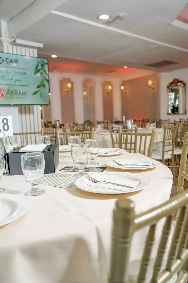 A table for ten set with white linens and gold chiavari chairs, sponsor screen behind it