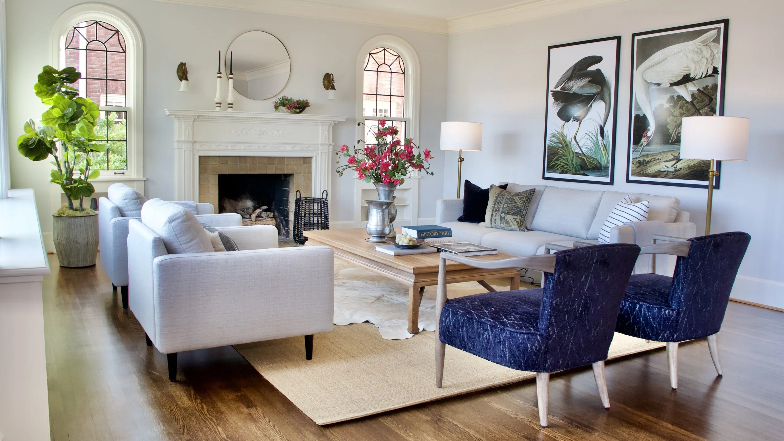 Pale sofas and deep blue chairs gathered around a wood table, with flowering stems, large bird prints and arched windows.
