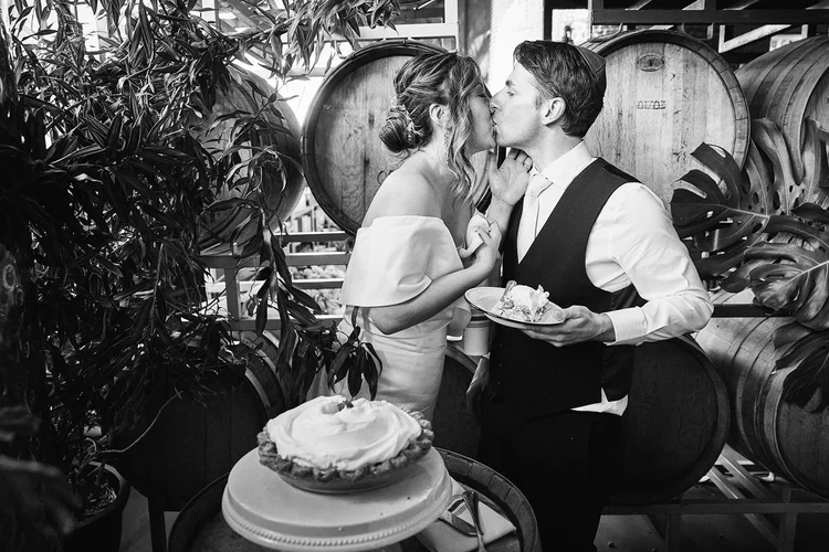 A bride and a groom kiss on their wedding day. The groom is holding a slice of pie.