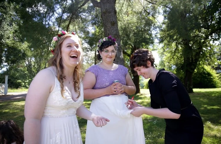 A wedding coordinator tries to get ice cream out of a bride's dress as her wife looks on.