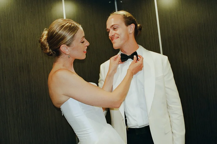 A bride and groom in an elevator on their wedding day. The bride is fixing the groom's bowtie.