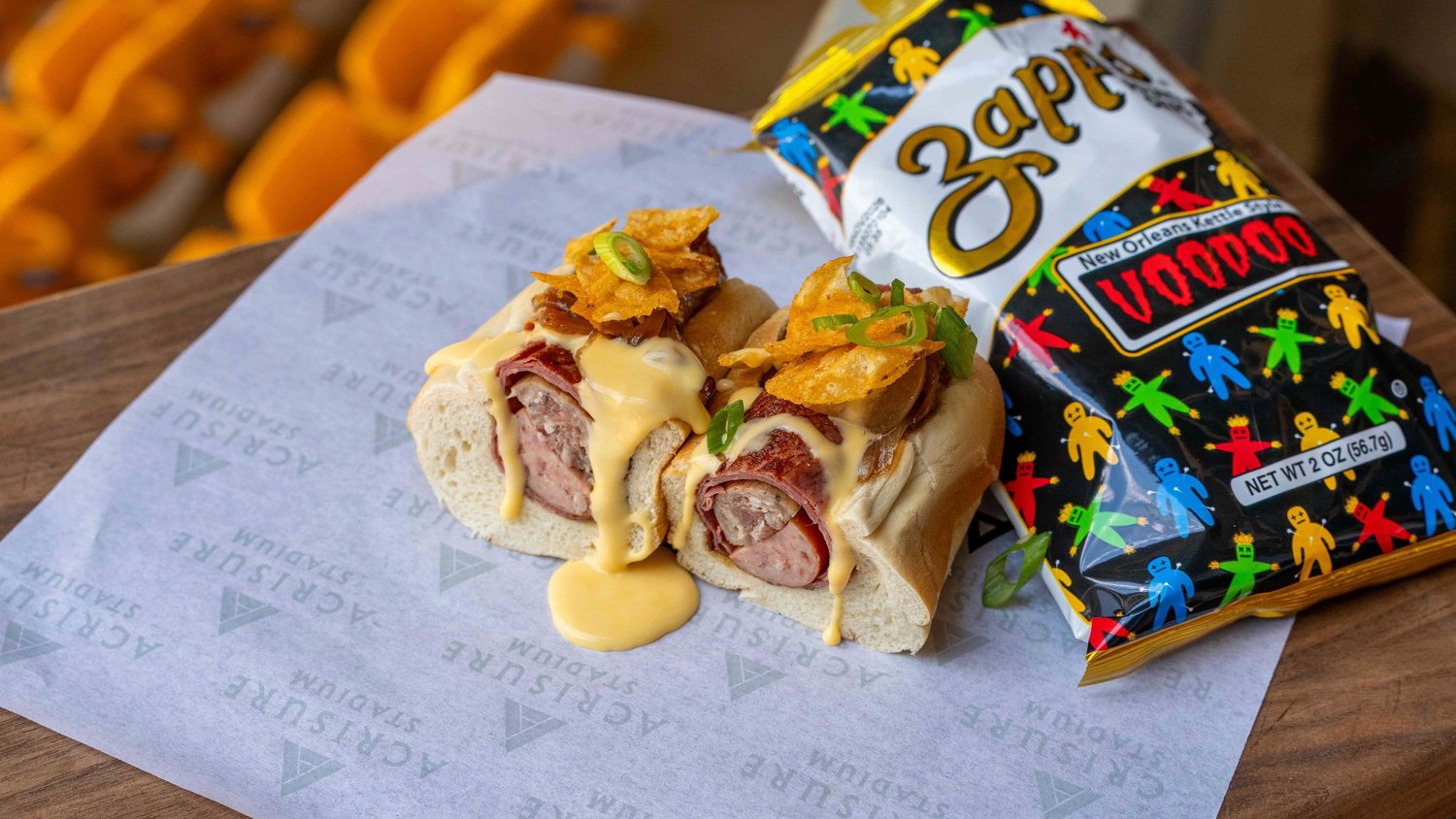 Two stadium sausage sandwiches covered in melted cheese sauce and topped with kettle chips and scallions beside a bag of Zapp's Voodoo chips, photographed by Ian Jones of ISJDESIGNS in Pittsburgh for Aramark Sports + Entertainment's Acrisure Stadium concessions client feature.