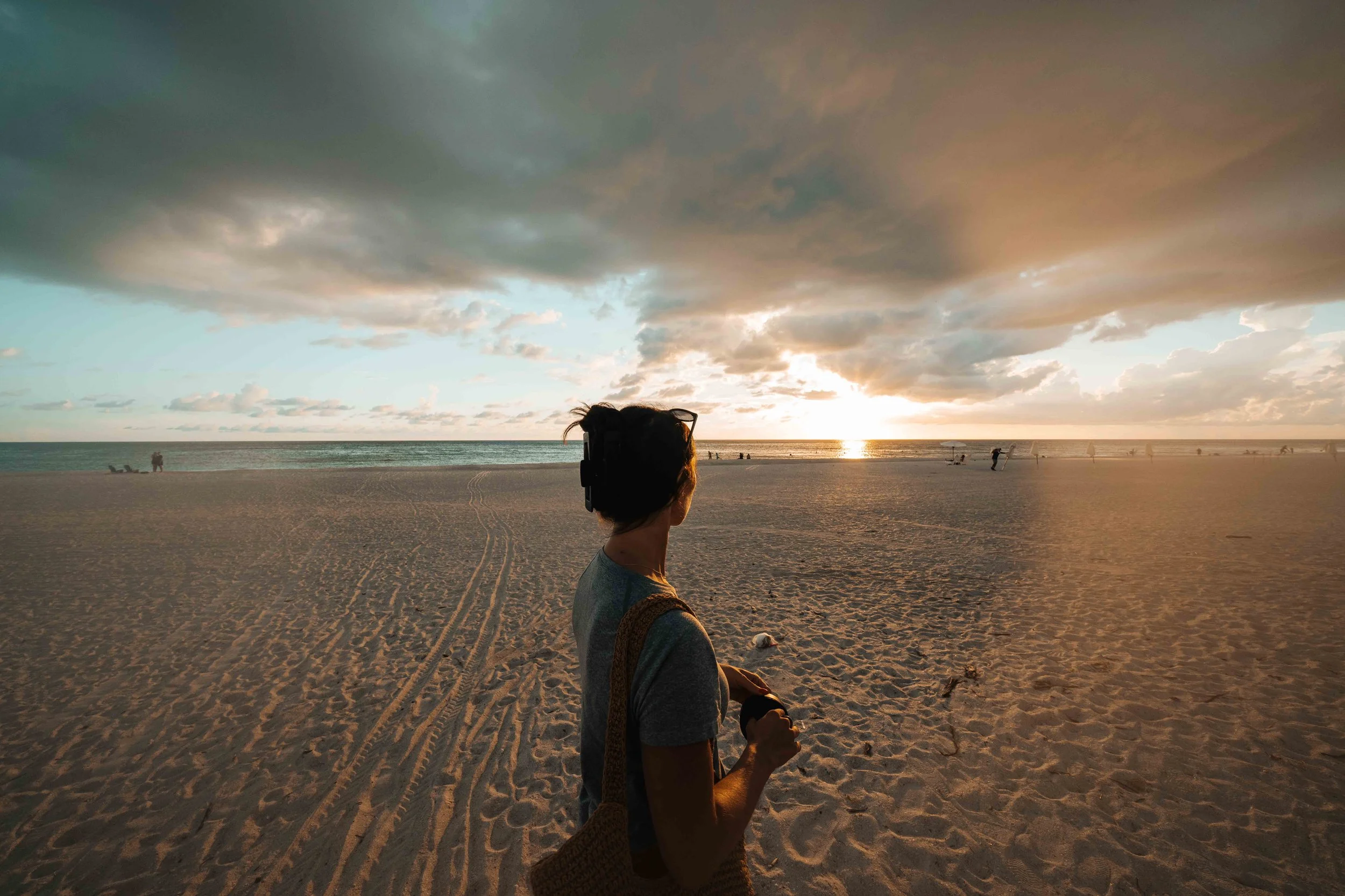 Woman standing on a wide Anna Maria Island beach watching the sunset, with the sun in frame and heavy clouds stretching across the sky