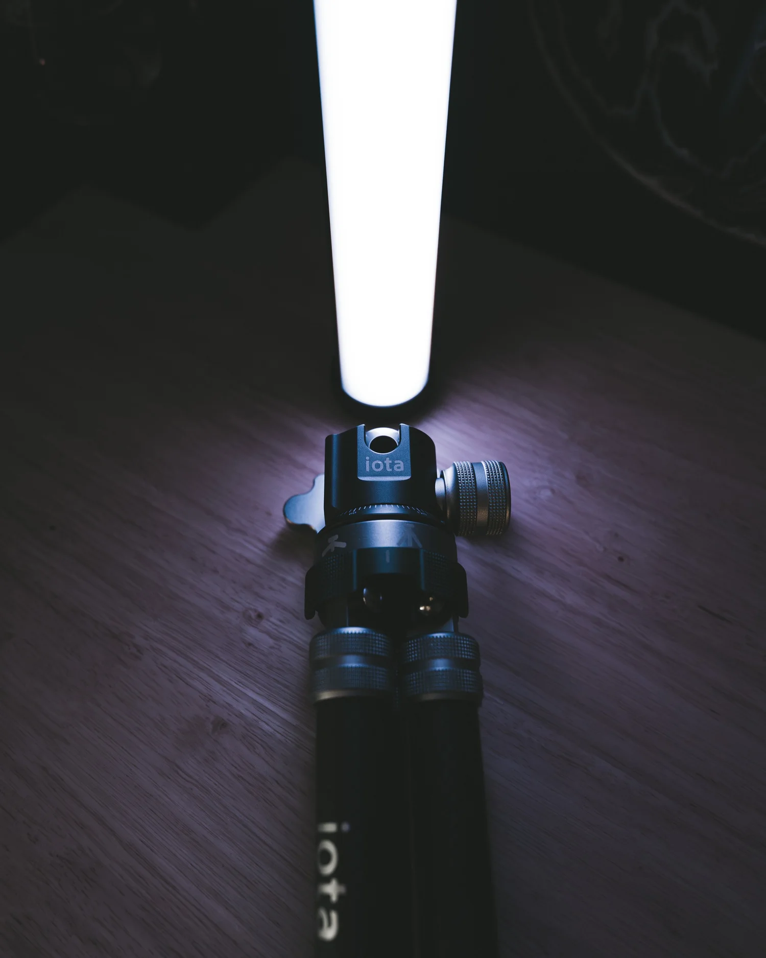 ProMaster iota tripod ballhead holding a glowing LED tube light, shot low and moody on a wood surface to show the product detail in use