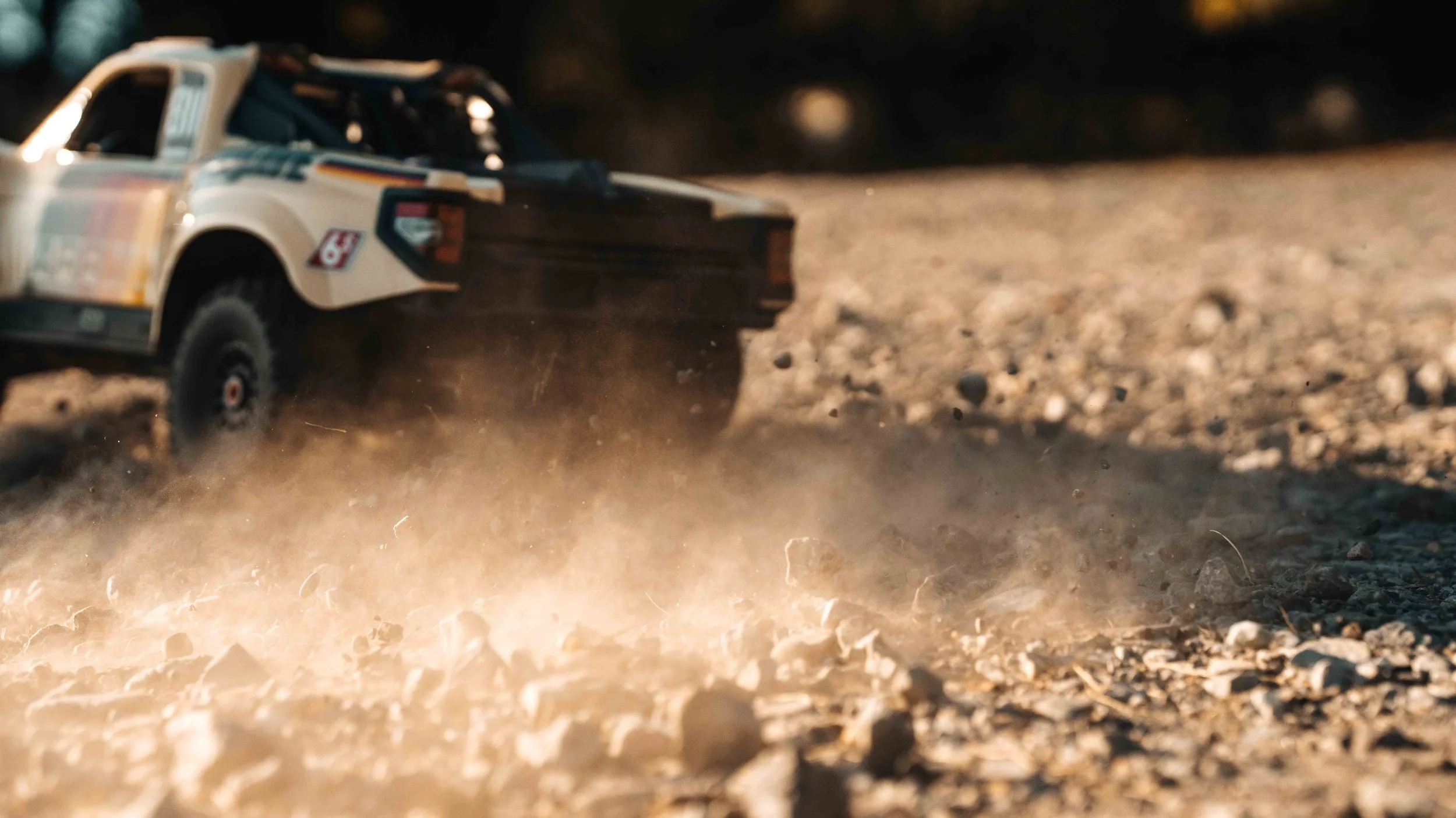 ARRMA Mojave 6S V3 RC desert truck accelerating across a gravel clearing, kicking up a bright spray of dust and loose stones behind the rear tires.