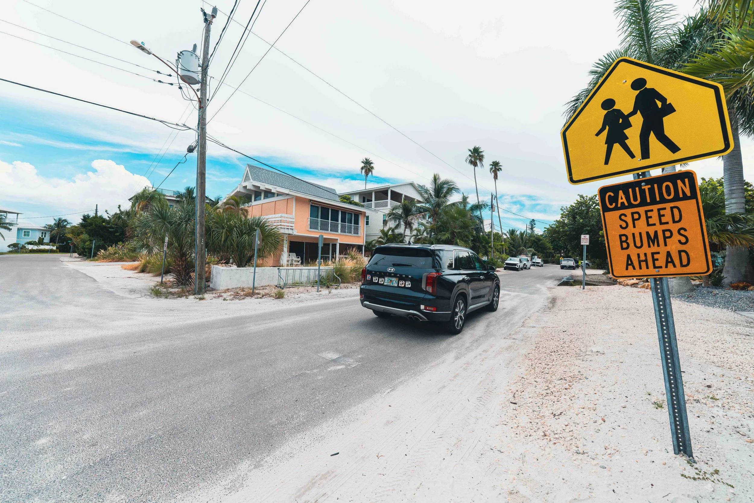 Anna Maria Island side street with a pedestrian crossing sign and a caution speed bumps ahead sign up front, an SUV, palms and beach houses behind