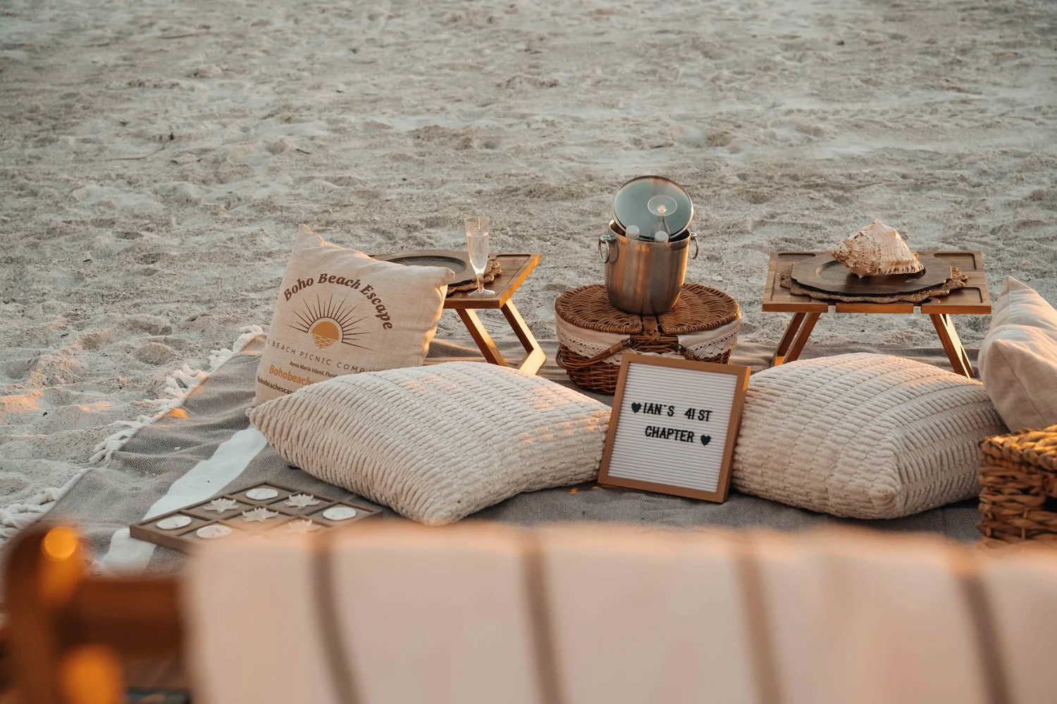 Beach picnic with low wooden tables, ice bucket, cushions, and celebration details, hospitality lifestyle photography for booking pages and experience marketing.