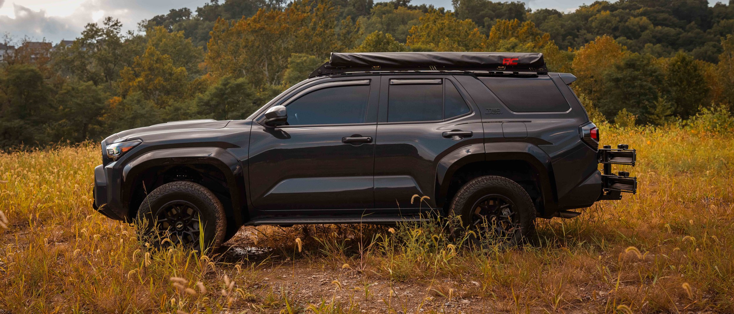 Full side profile of Ian Jones' 2025 Toyota 4Runner TRD Sport in Underground gray parked in a fall field, showing the Cali Raised LED Premium Roof Rack, Rough Country retractable awning, gloss black wide fender flares, RockTrix RT107 wheels, Cooper all-terrain tires, and a rear hitch-mounted bike rack.