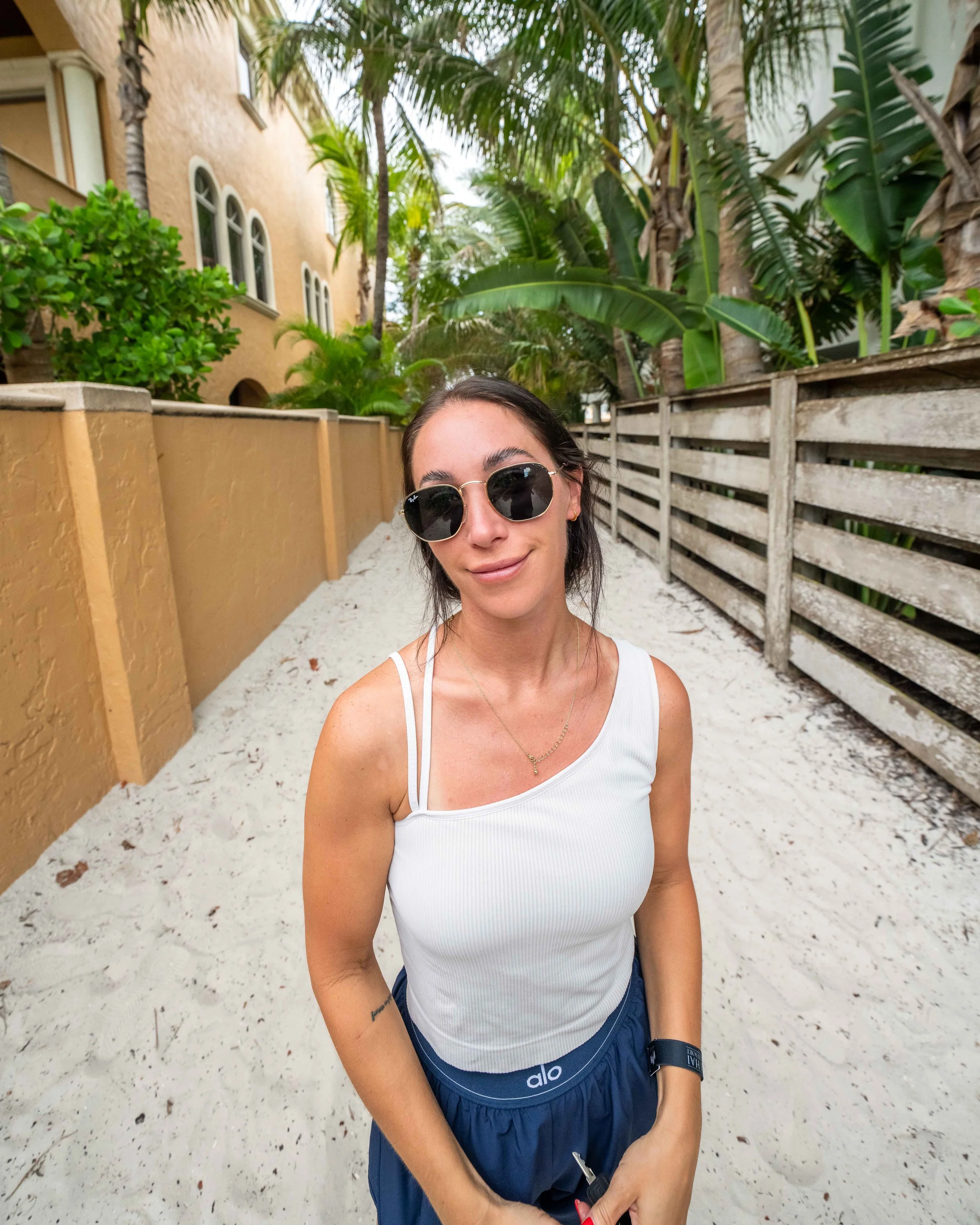 Environmental portrait of a smiling woman in sunglasses and a white tank top on a sandy path between a stucco wall and a wooden fence, palms overhead