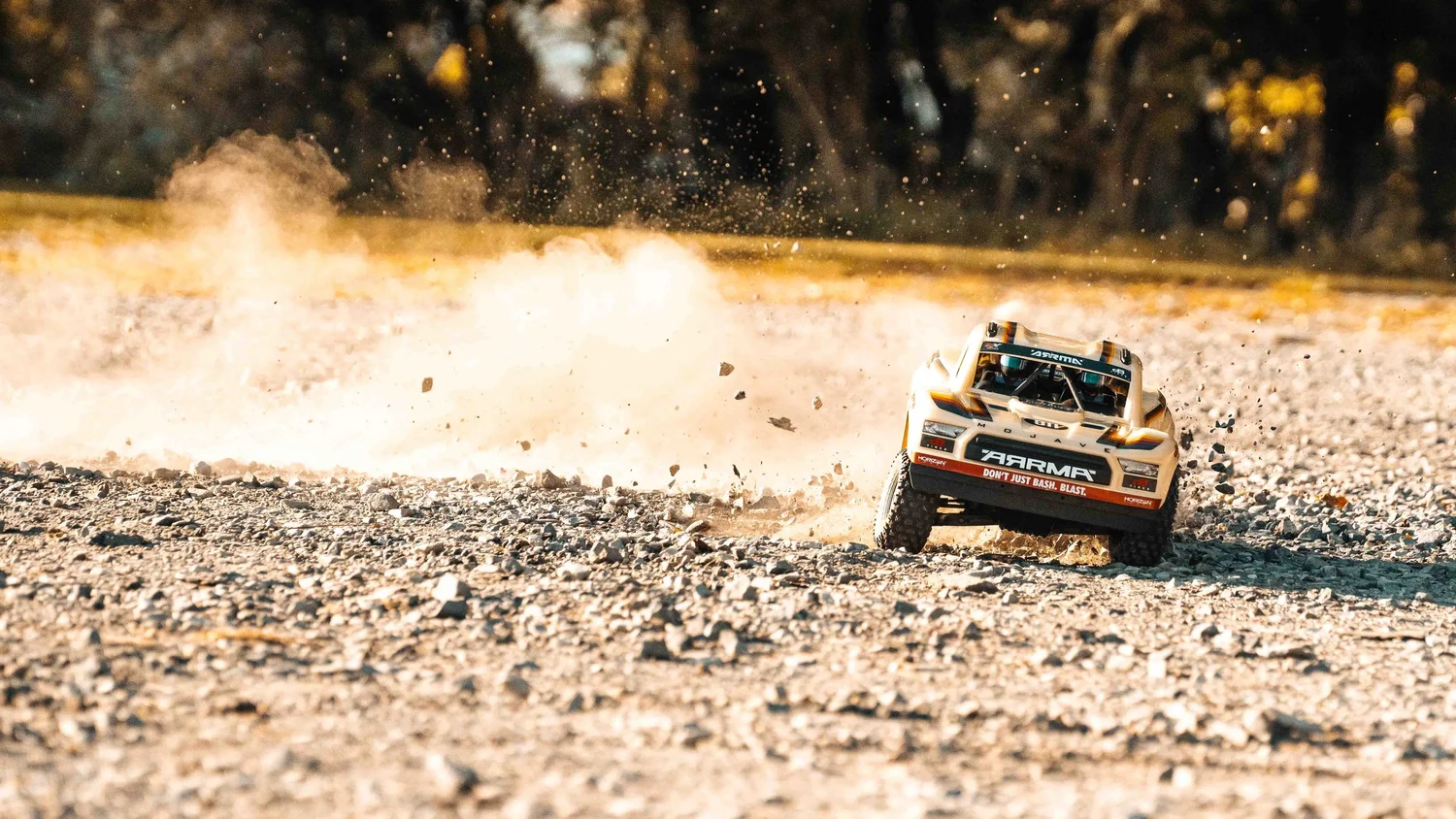 Front-facing ARRMA Mojave 6S V3 remote-control desert truck racing over loose gravel as a large, backlit dust cloud expands behind it.