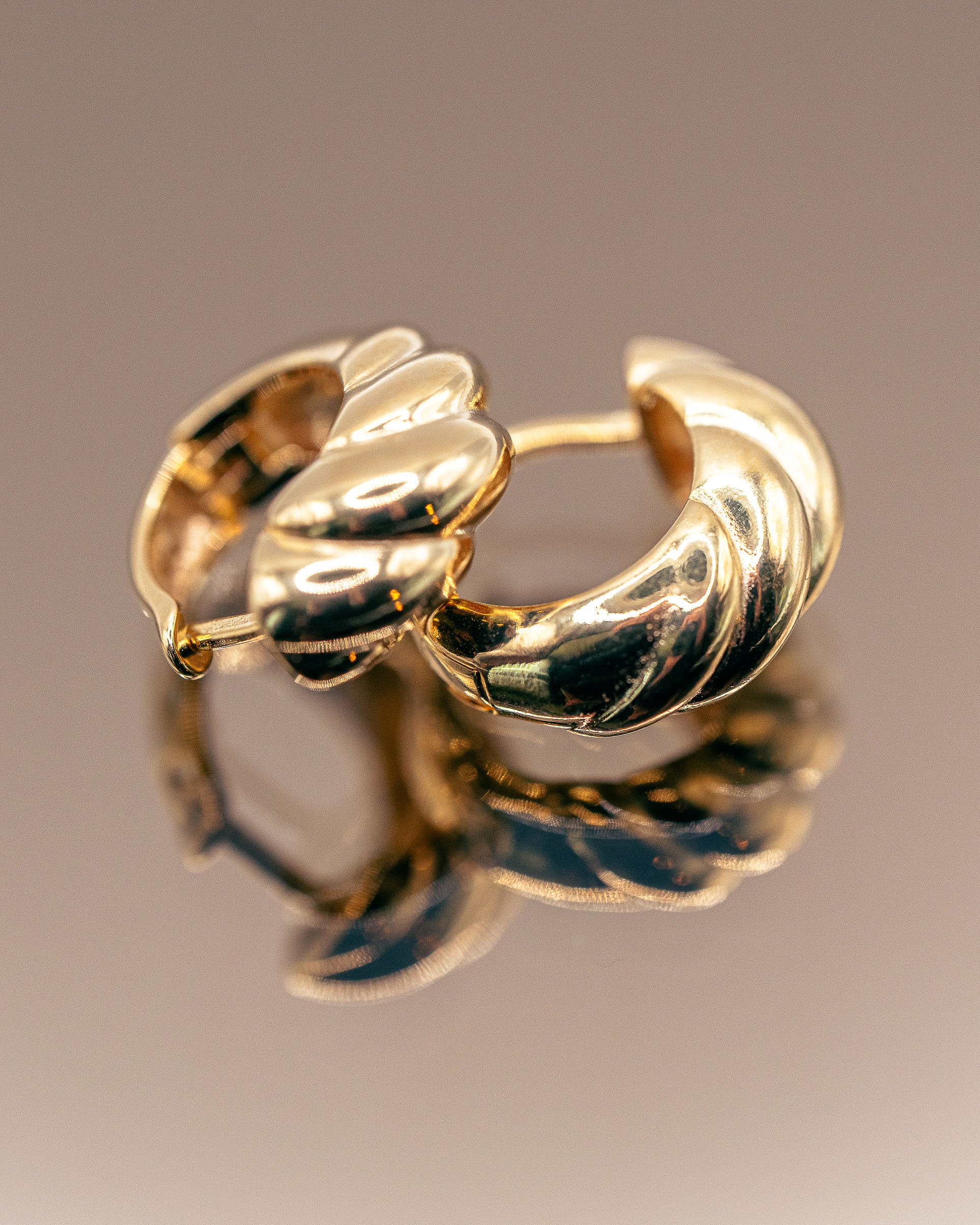 Gold croissant hoop earrings on a mirrored surface, polished metal lit to hold every curve. Ian Jones, ISJDESIGNS.