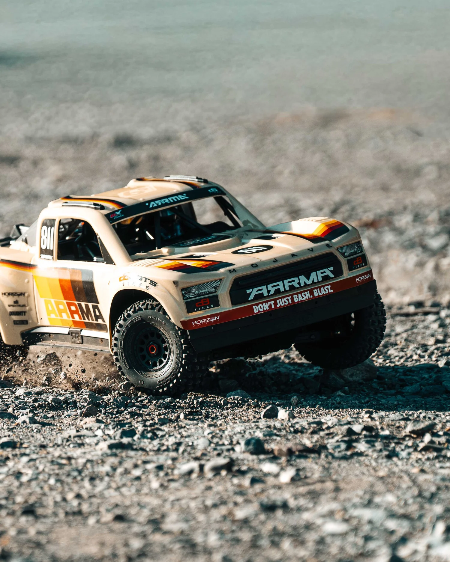 Front three-quarter view of the ARRMA Mojave 6S V3 desert truck on a gravel surface, showcasing its front grille, detailed racing graphics and knobby tires.