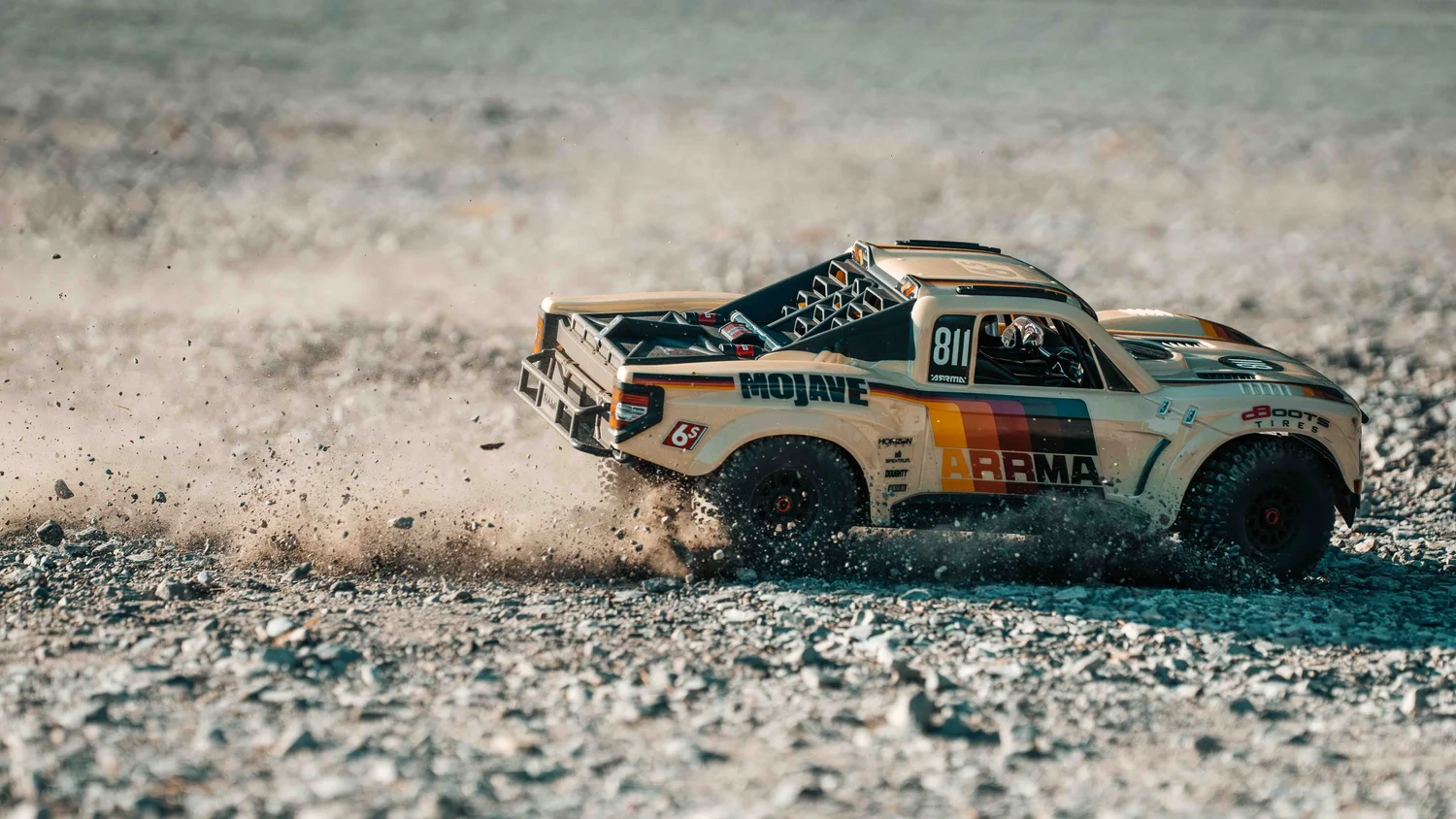 Side profile of a beige ARRMA Mojave 6S V3 RC truck sliding across rocky ground, with a trail of dust behind the body and tires.