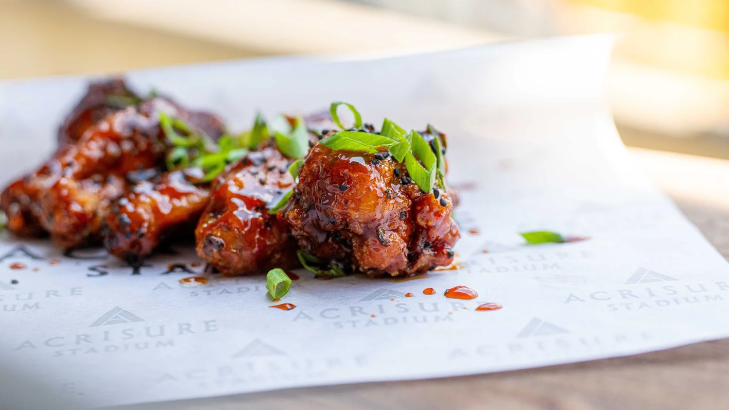 Close-up of sticky glazed chicken wings finished with black sesame seeds and sliced scallions on Acrisure Stadium-branded paper, photographed by Ian Jones of ISJDESIGNS in Pittsburgh for Aramark Sports + Entertainment, an ISJDESIGNS food photography client.