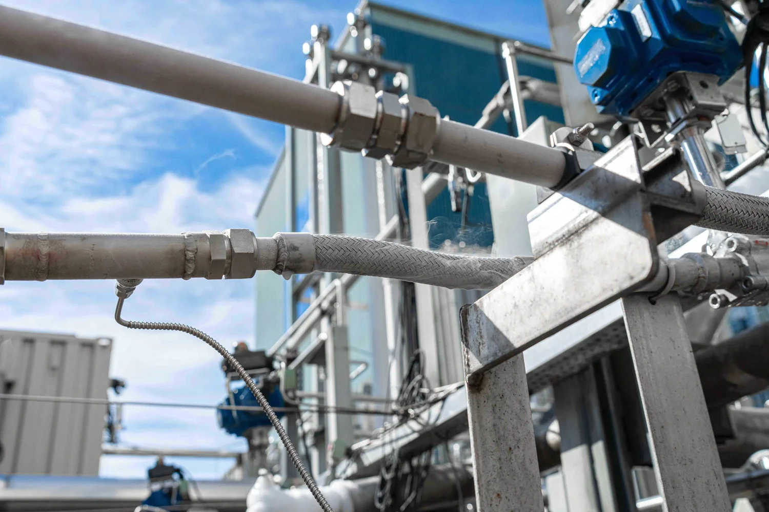 Industrial process photography of a cryogenic transfer line, braided hose, actuated valve and vaporizer equipment, documenting technical infrastructure inside a manufacturing facility.