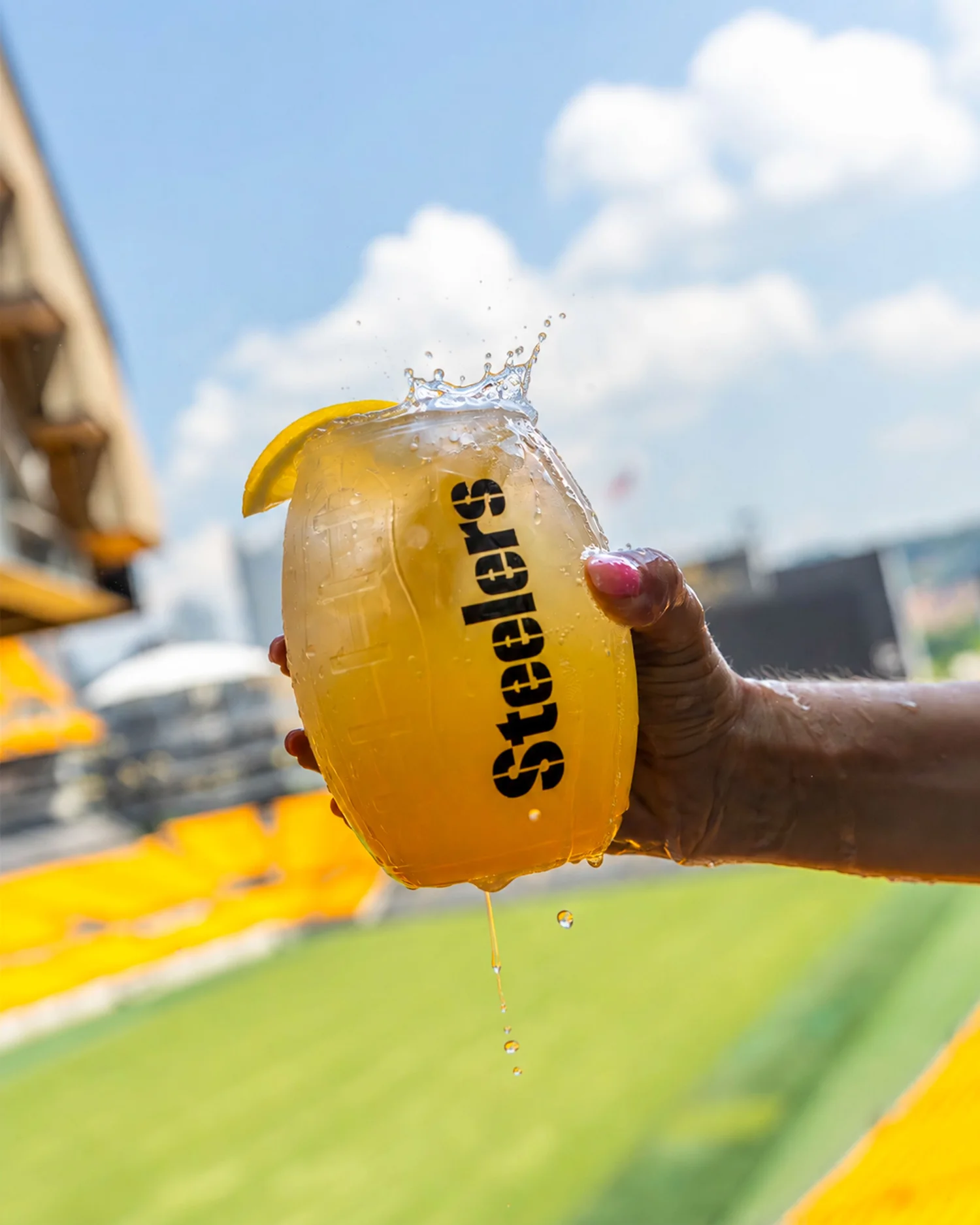 The Immaculate Reception cocktail splashes from a Steelers-branded glass held above the field at Acrisure Stadium, photographed by Ian Jones of ISJDESIGNS in Pittsburgh for Aramark Sports + Entertainment, an ISJDESIGNS food and beverage photography client.