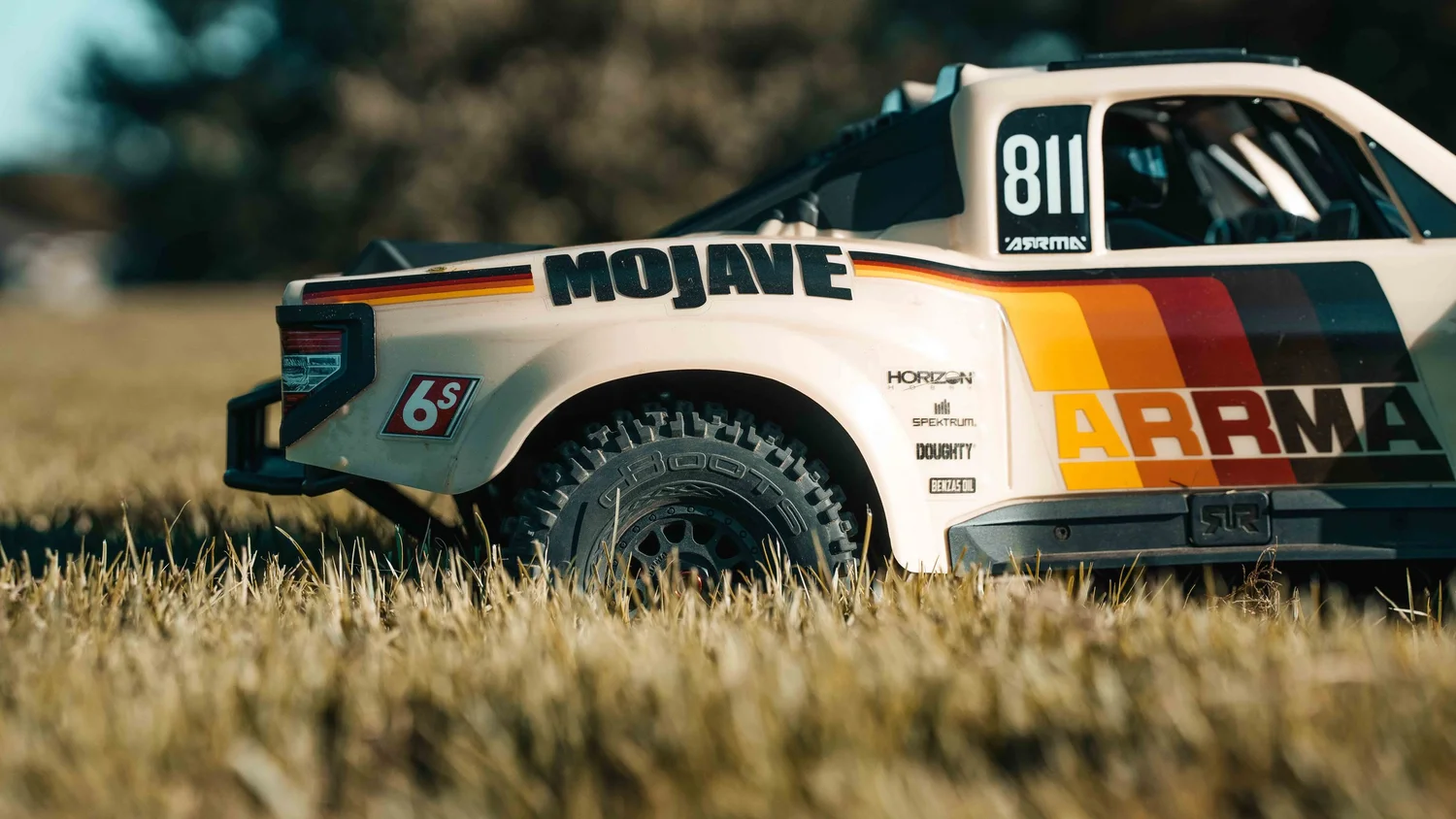 Close side view of a beige and orange ARRMA Mojave 6S V3 RC desert truck parked in grass, highlighting its racing livery, wheel, fender and realistic truck body.