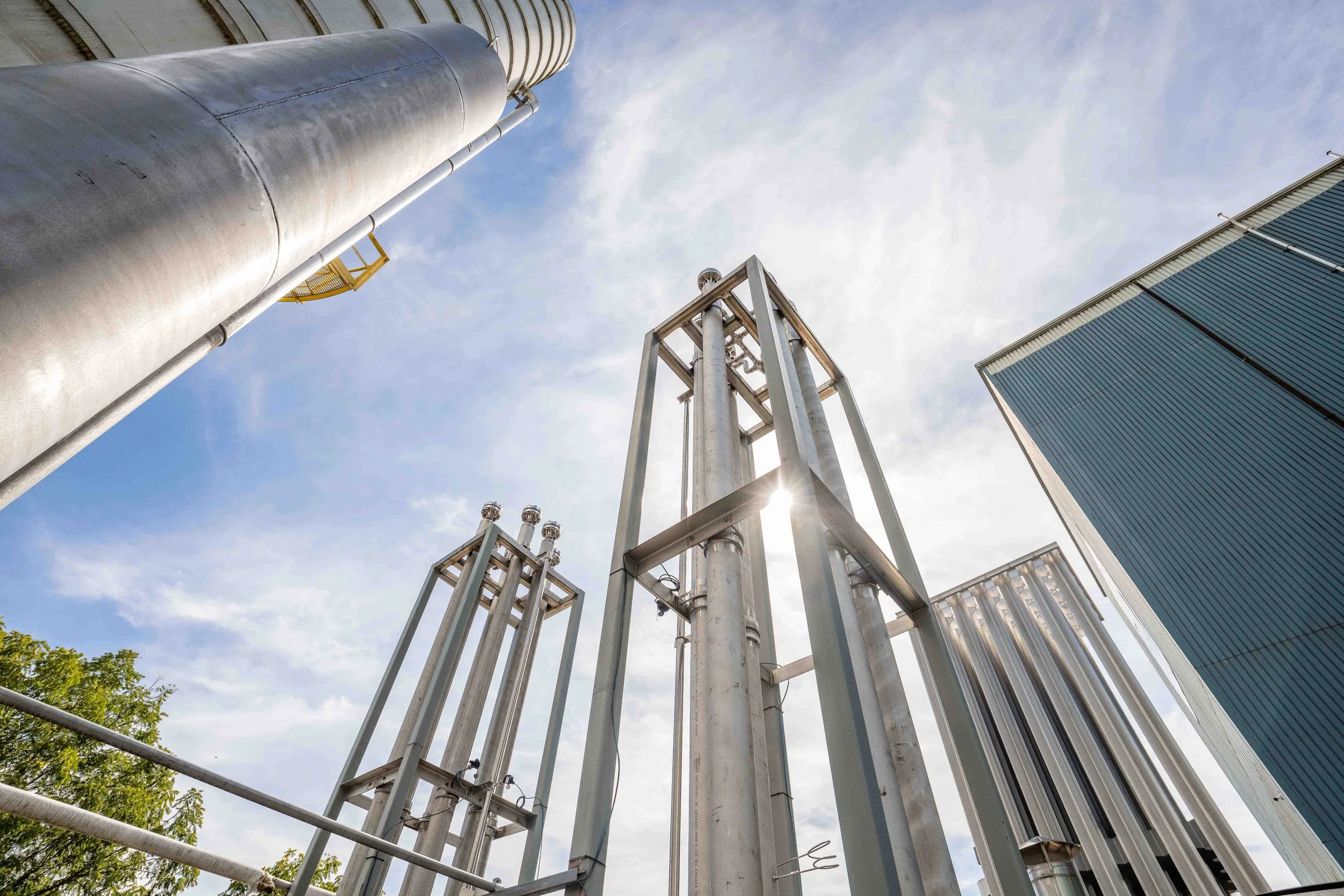 Wide low-angle industrial photography of cryogenic vaporizers, a storage silo and manufacturing building, capturing the scale of process equipment and industrial plant infrastructure.