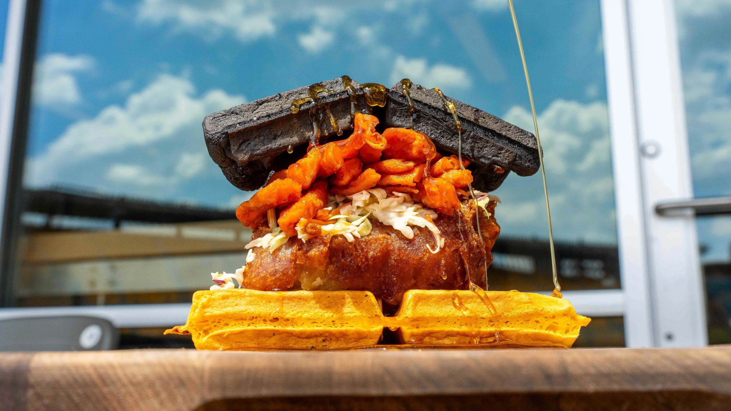 Fried chicken and waffle stadium sandwich layered with slaw and spicy cheese curls, finished with syrup dripping over dark and golden waffles, photographed by Ian Jones of ISJDESIGNS in Pittsburgh for Aramark Sports + Entertainment's Acrisure Stadium client feature.