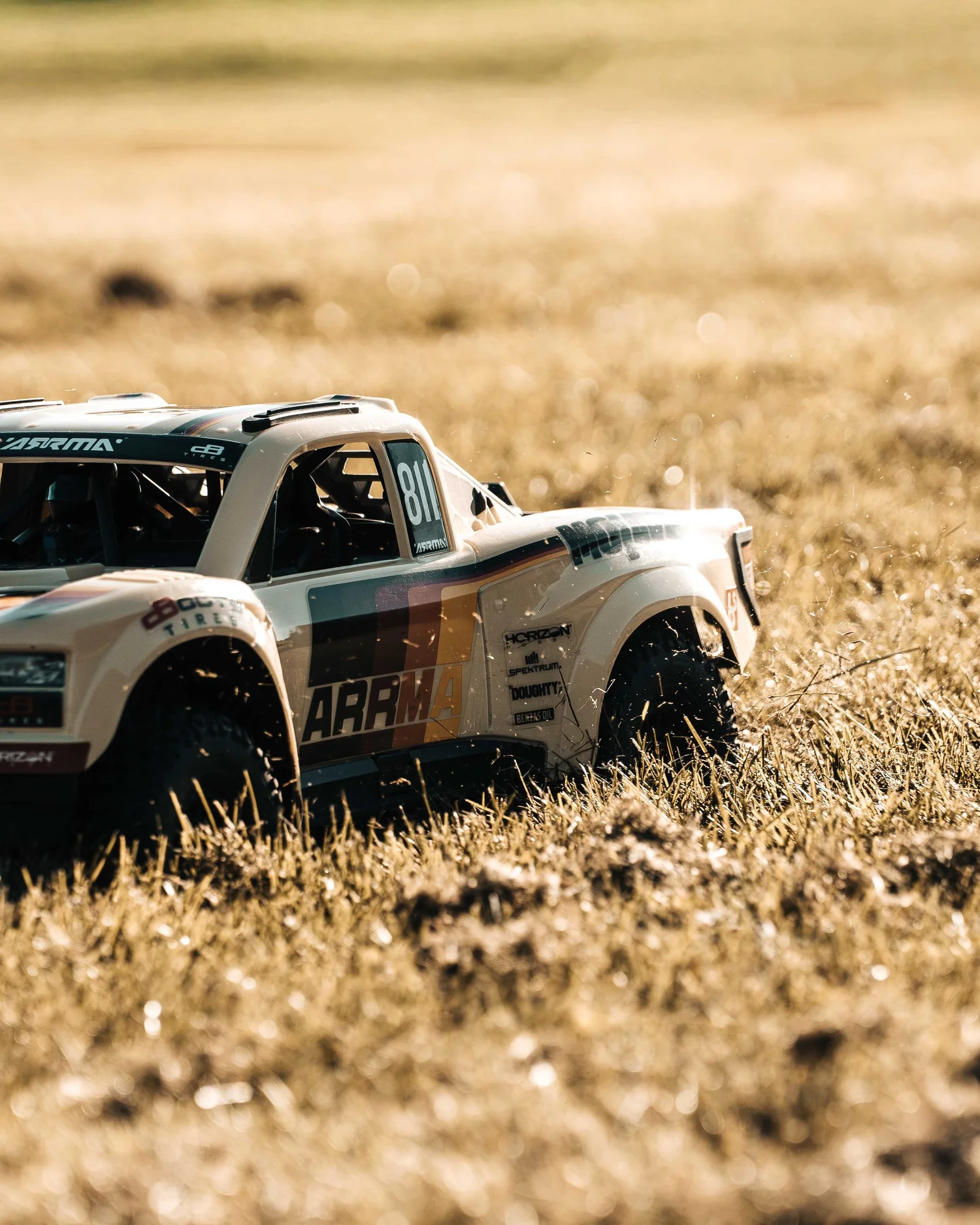 ARRMA Mojave 6S V3 remote-control desert truck in a dry grassy field, photographed from a front three-quarter angle with its beige racing body and roll cage visible.