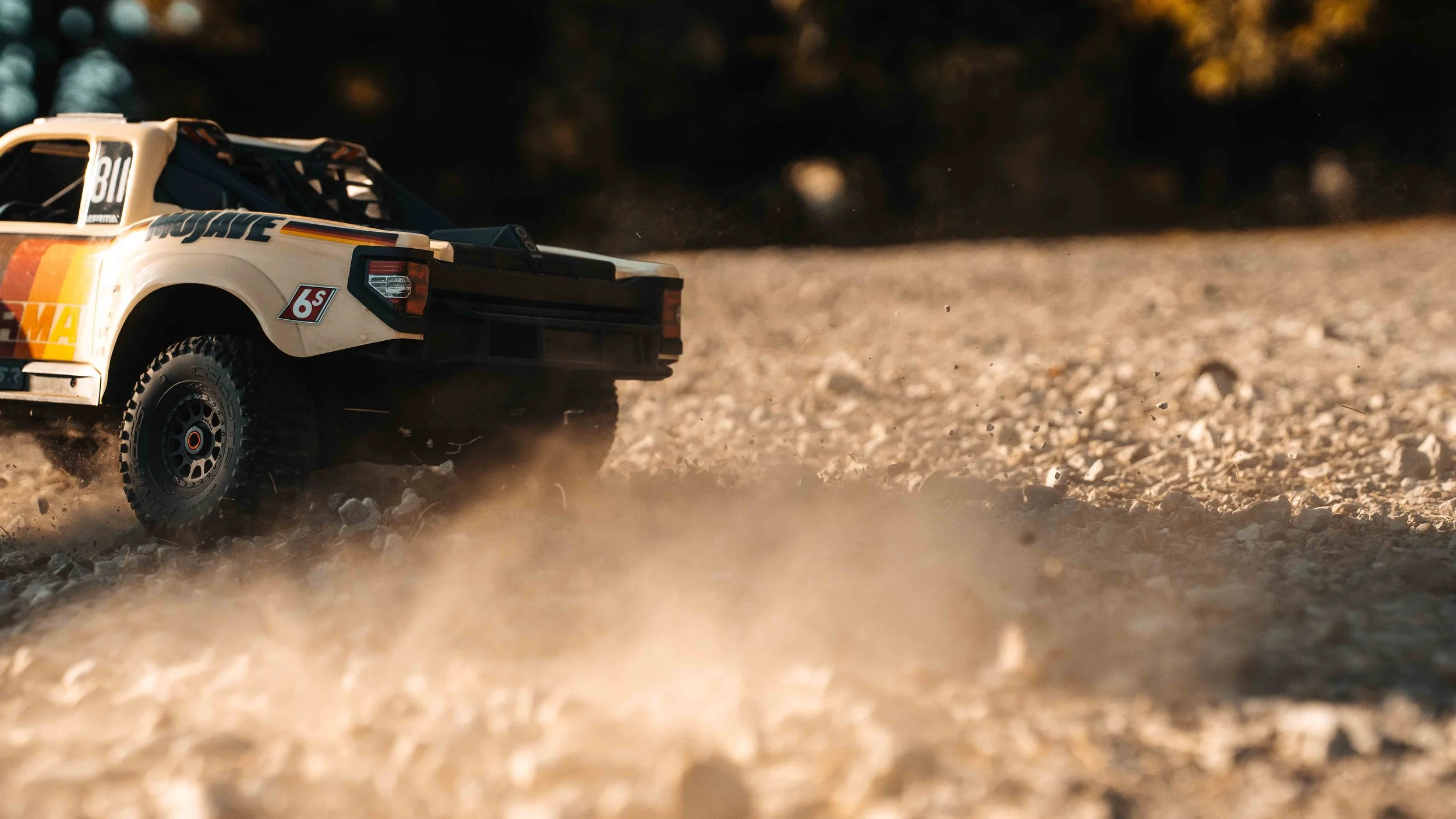 Close rear three-quarter view of the ARRMA Mojave 6S V3 off-road RC truck spraying gravel and dust as it speeds over rough ground in golden light.