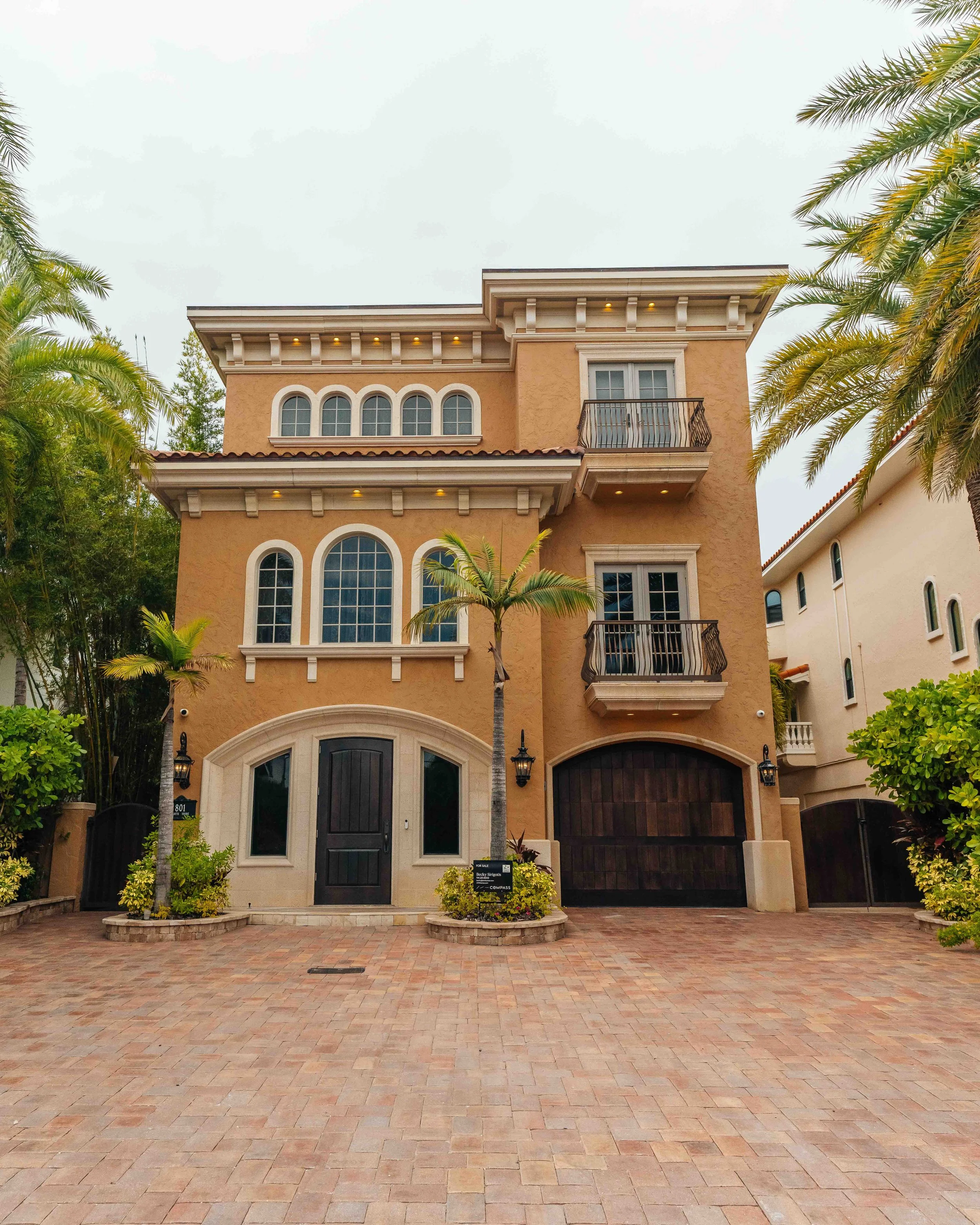 Vertical frame of a three-story Mediterranean-style home with arched windows, balconies and a brick-paver driveway, flanked by tall palms