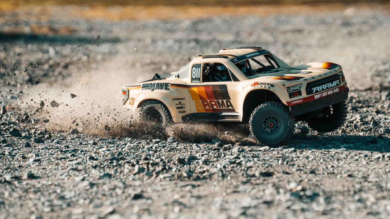 Low side-angle action photograph of a beige ARRMA Mojave 6S V3 desert truck moving across a rocky surface, its suspension and large off-road tires visible.