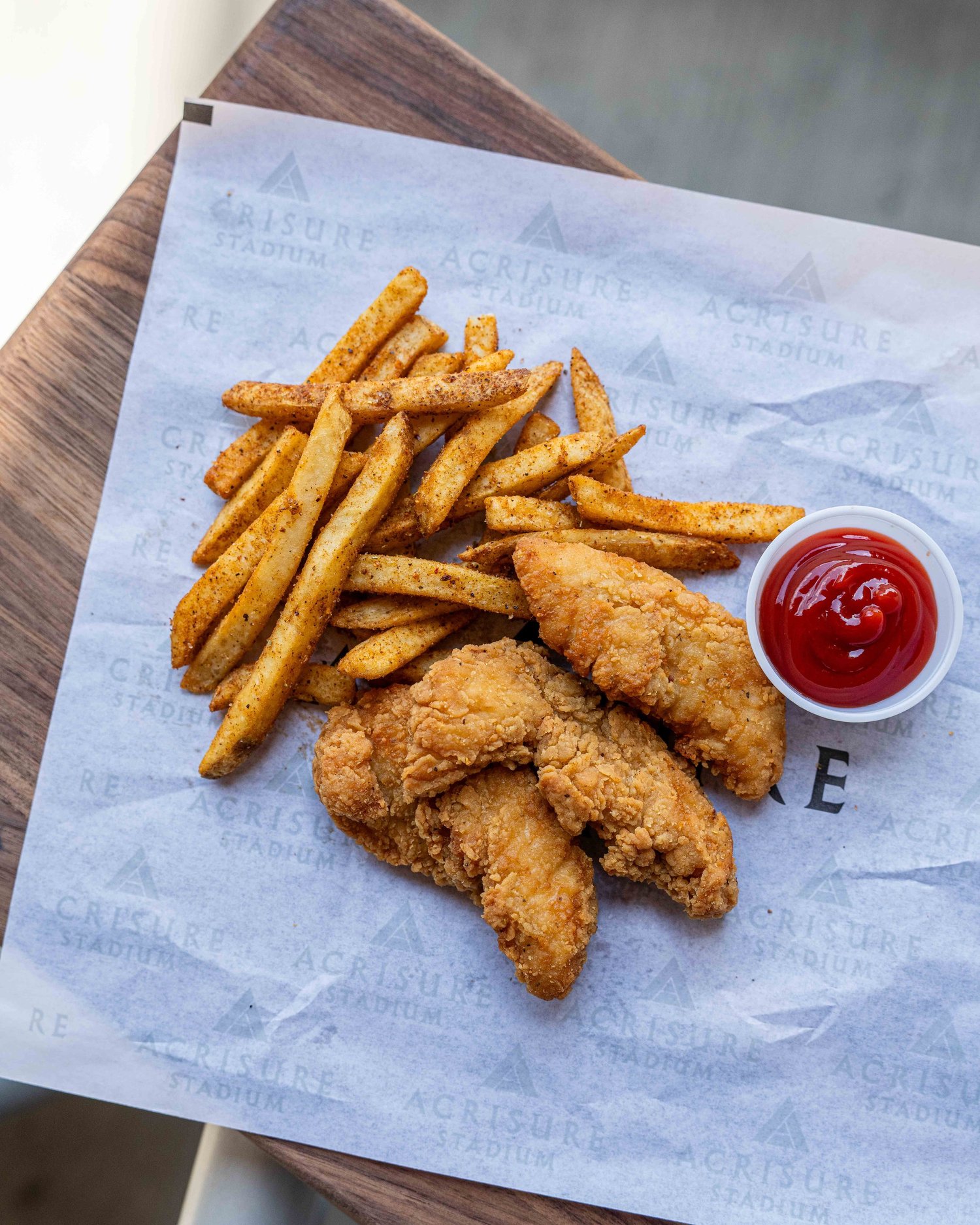 Crispy chicken tenders served with seasoned French fries and a cup of ketchup on Acrisure Stadium-branded paper, photographed by Ian Jones of ISJDESIGNS in Pittsburgh for Aramark Sports + Entertainment's stadium concessions client feature.