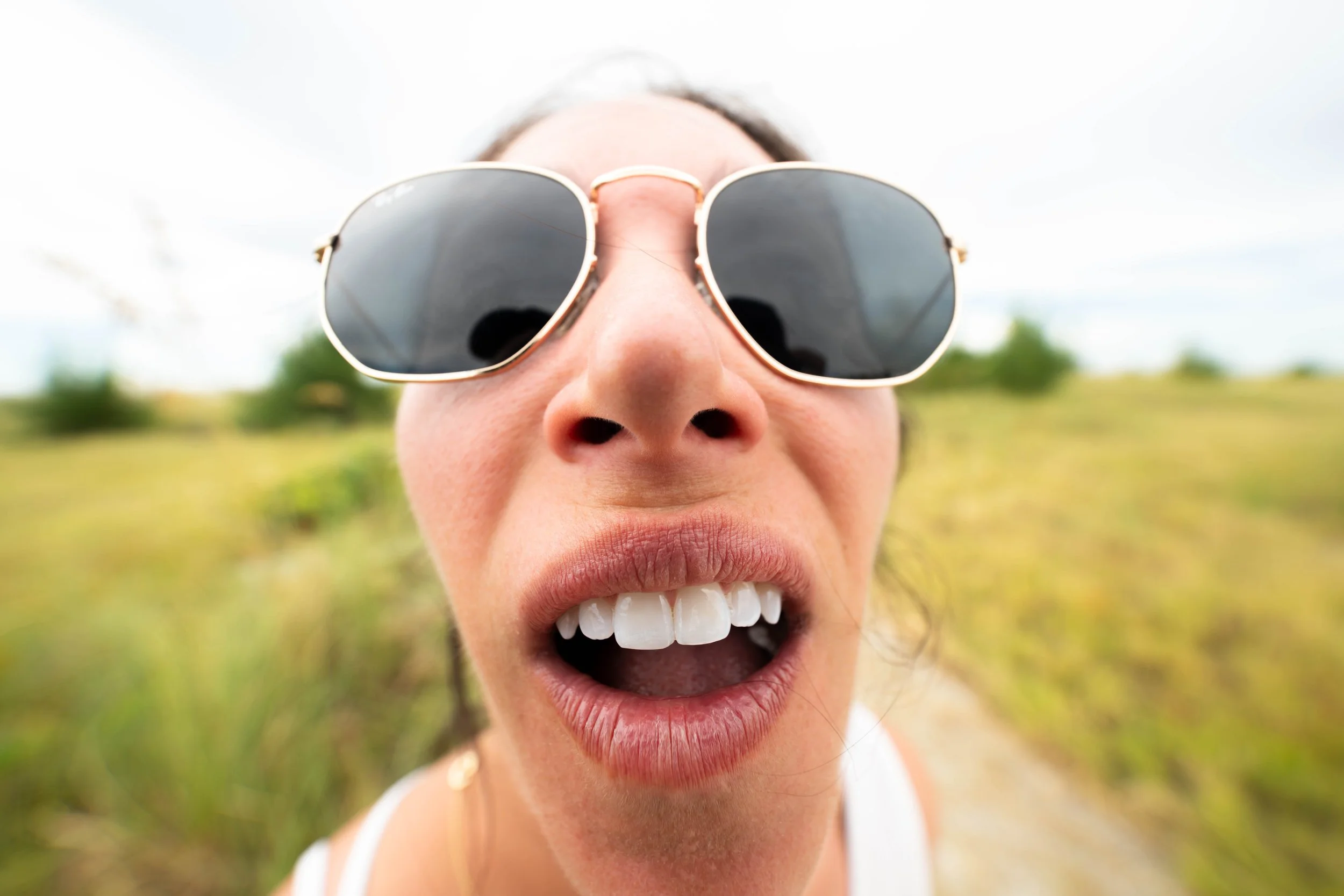 Playful ultra-wide close-up with the lens inches from a woman's face, her sunglasses and grin stretched large against green dune grass