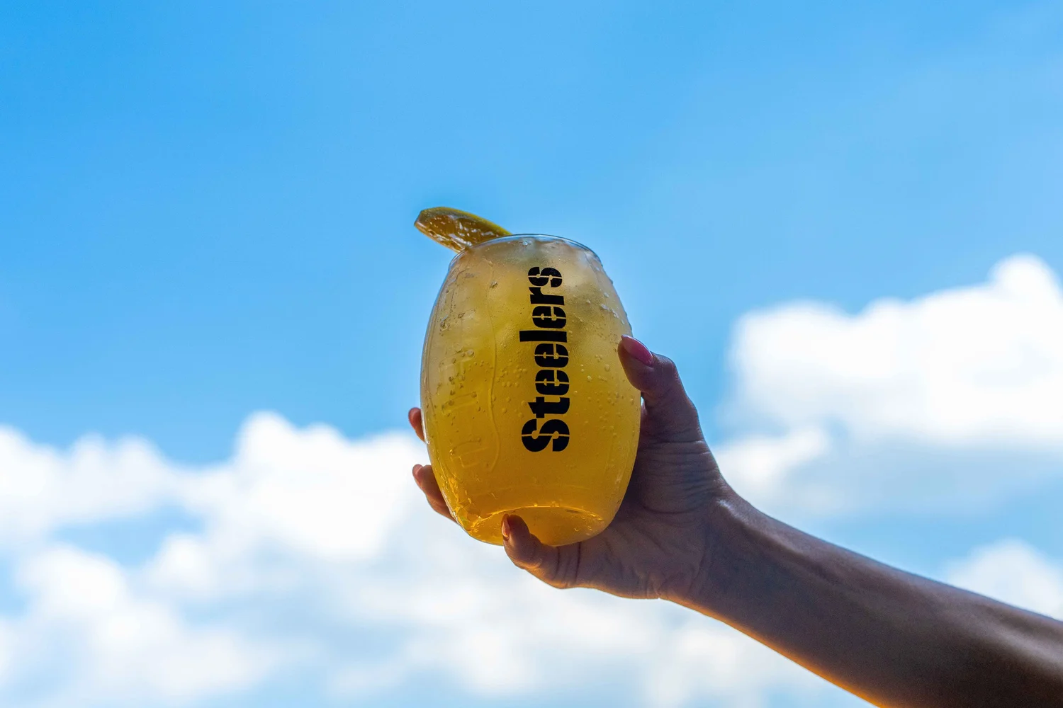 A hand raises a chilled golden cocktail in a Steelers-branded glass against a bright blue Pittsburgh sky, photographed by Ian Jones of ISJDESIGNS for Aramark Sports + Entertainment's Acrisure Stadium concessions client feature.