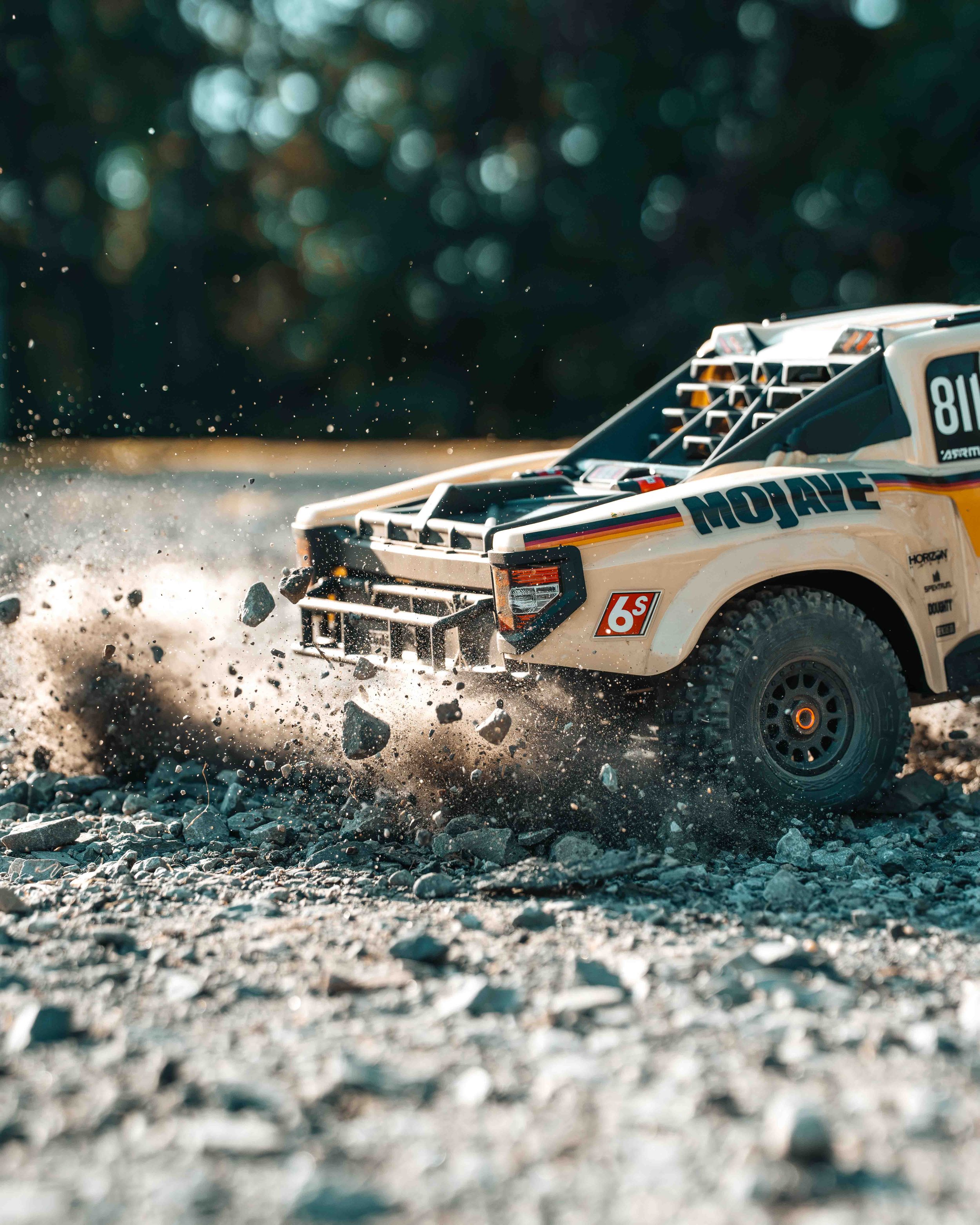 Low-angle close-up of the ARRMA Mojave 6S V3 desert truck charging through loose gravel, with stones flying past its front bumper and spinning tire.