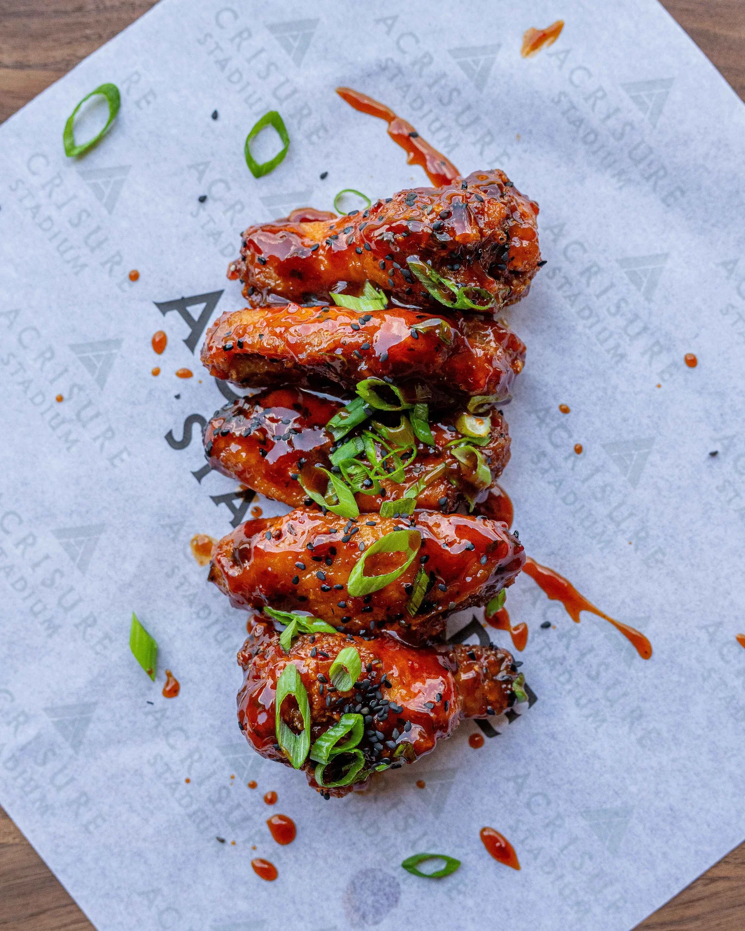 Overhead view of five glossy glazed chicken wings garnished with black sesame seeds and sliced scallions on Acrisure Stadium-branded paper, photographed by Ian Jones of ISJDESIGNS in Pittsburgh for Aramark Sports + Entertainment, an ISJDESIGNS food photography client.