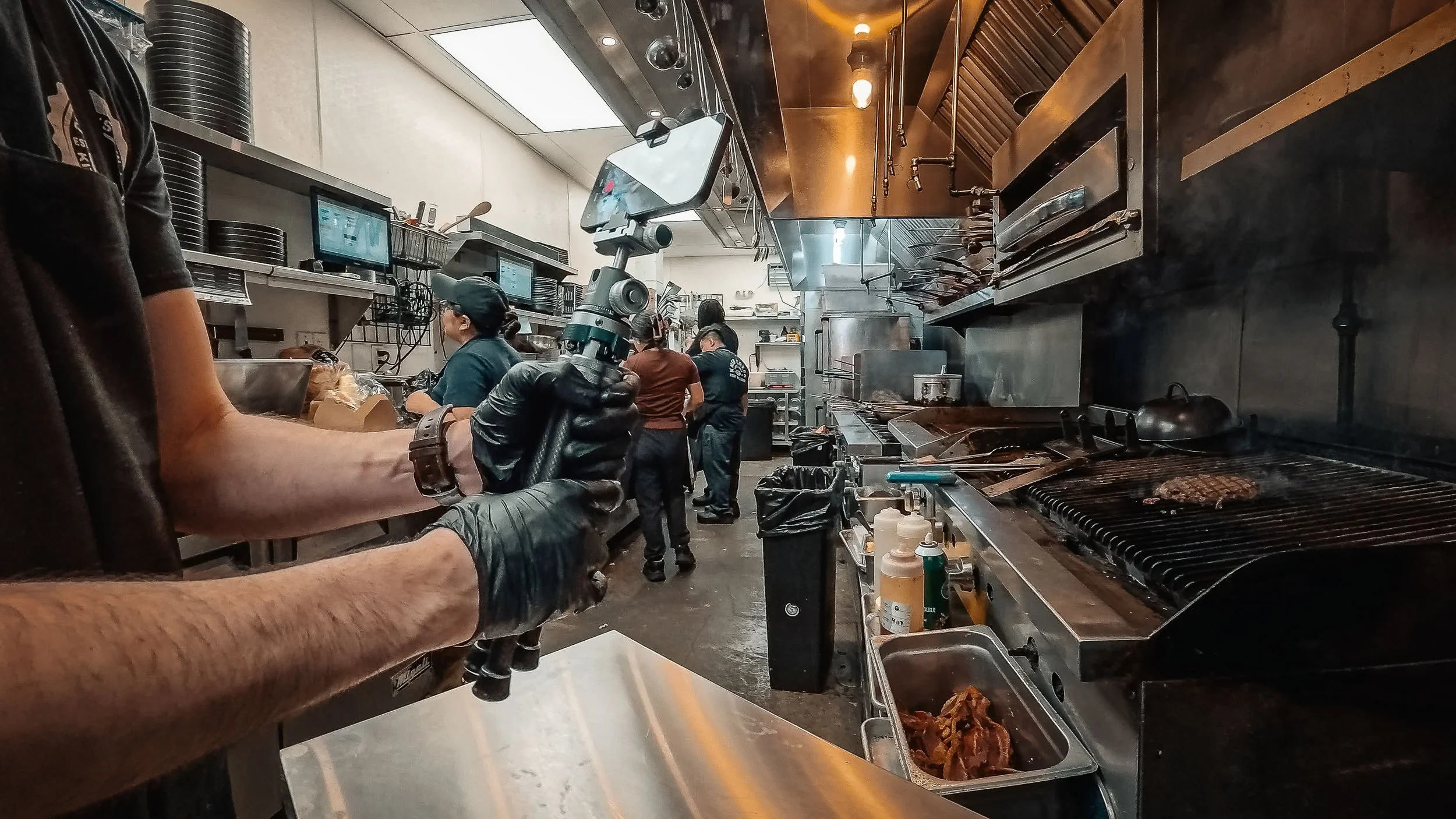 ProMaster iota 219 tripod holding an iPhone in video mode on a restaurant kitchen line while a gloved cook works the grill