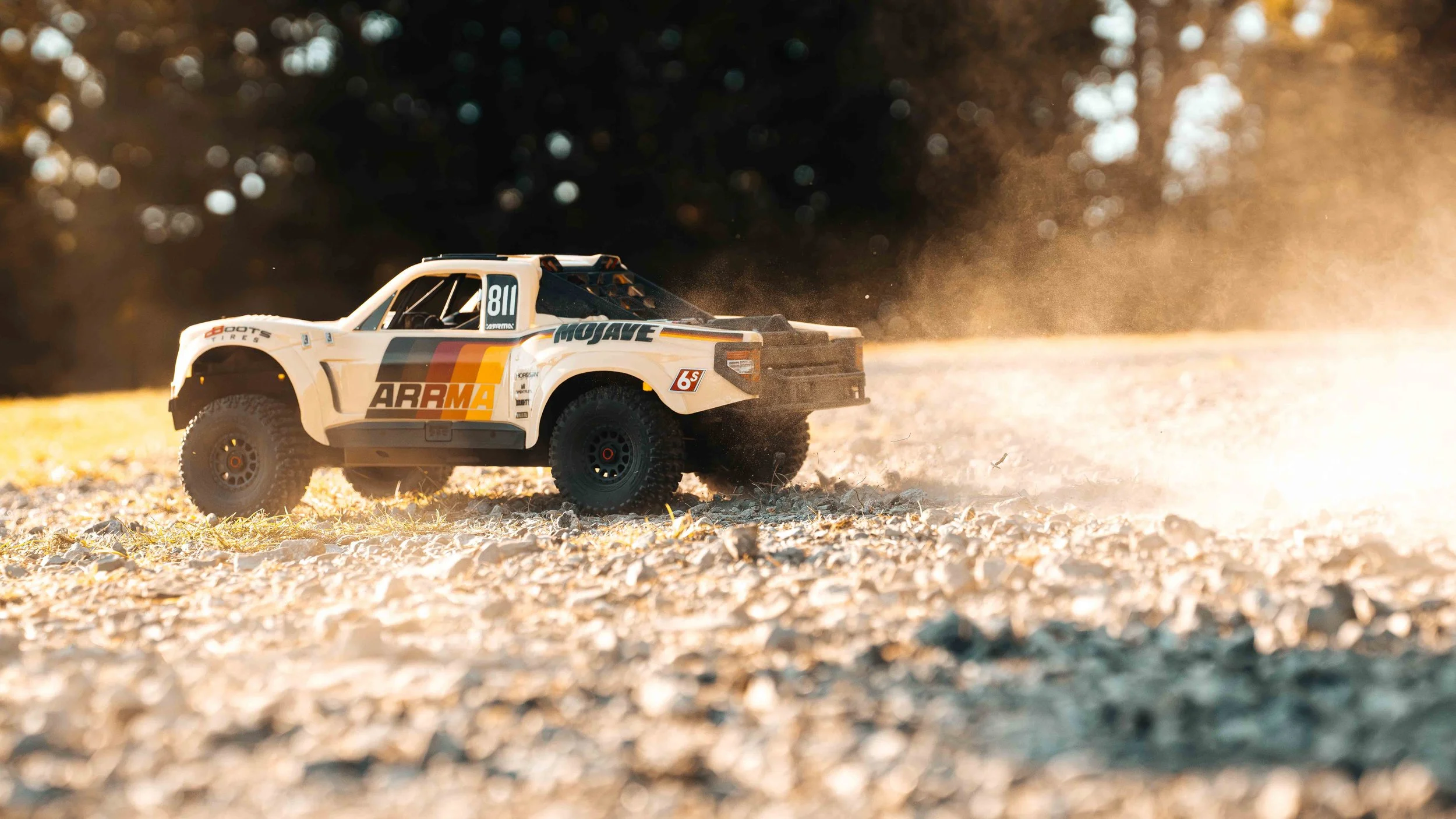 Sunlit action shot of the ARRMA Mojave 6S V3 RC truck driving across gravel, with a glowing cloud of dust trailing behind its beige and orange body.