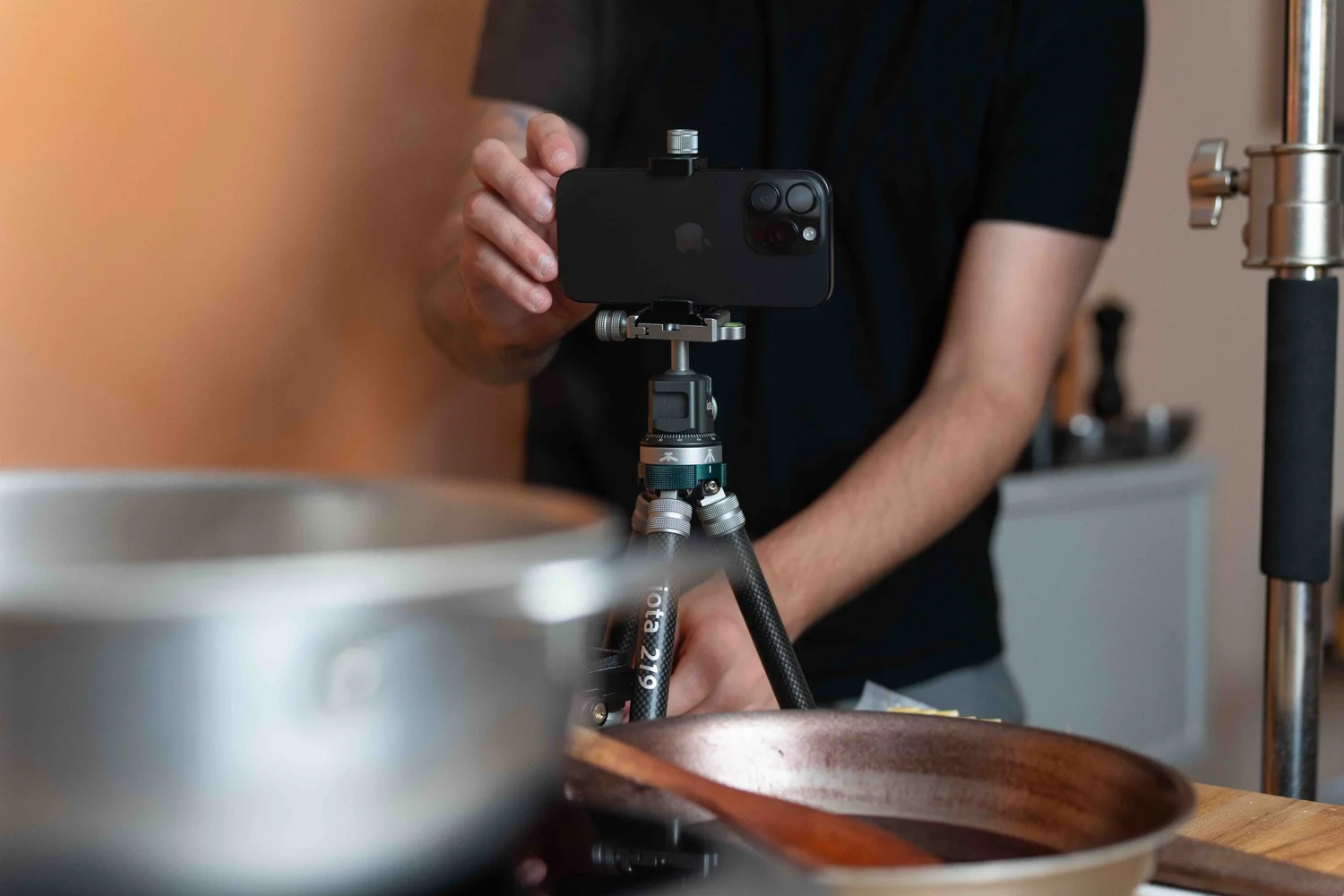 Hand adjusting an iPhone mounted on a ProMaster iota 219 tabletop tripod on a kitchen counter beside a saucepan and wooden bowl