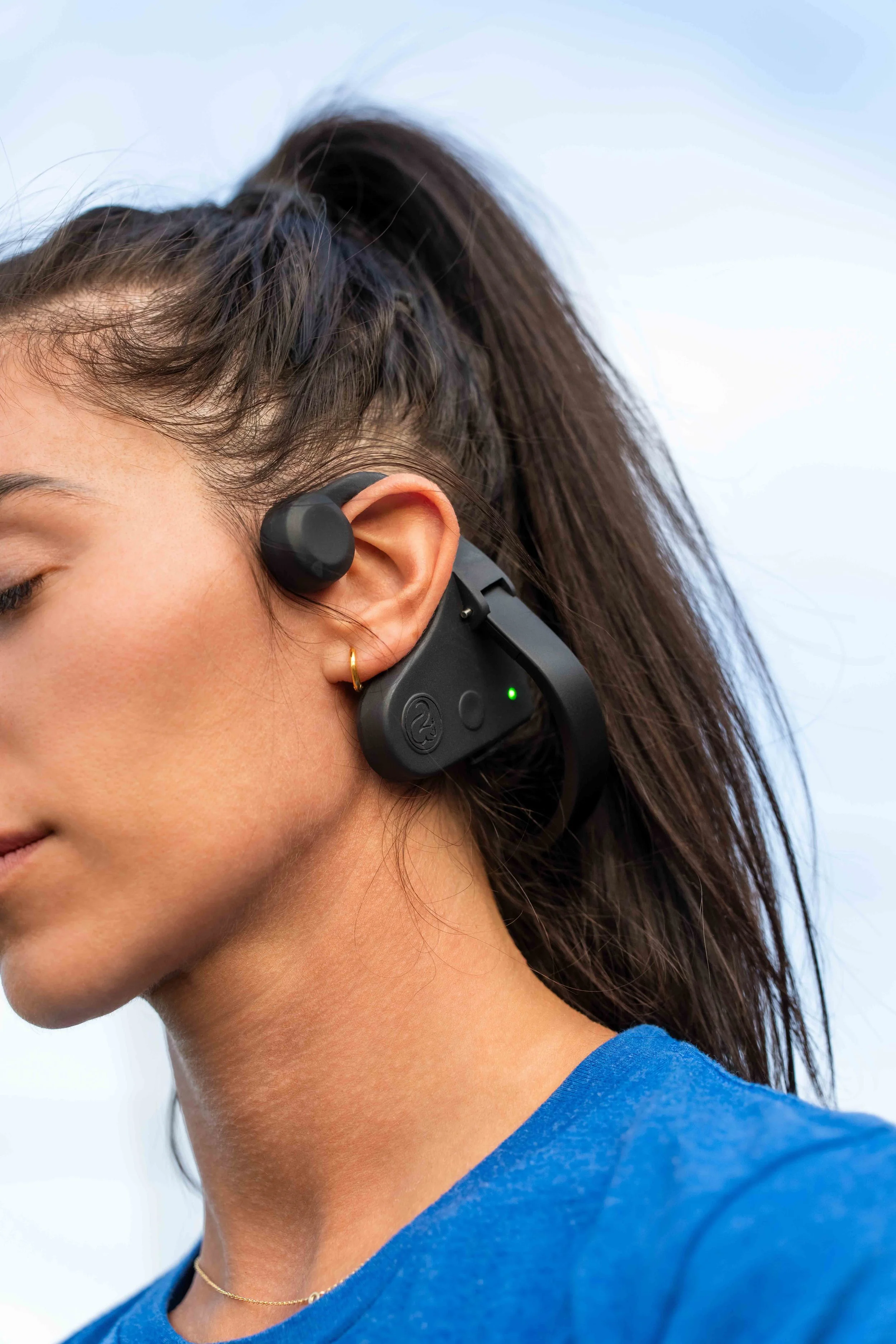 Lifestyle product photo of a model with a high ponytail wearing a black open-ear headset with a green status light, outdoors against a soft blue sky