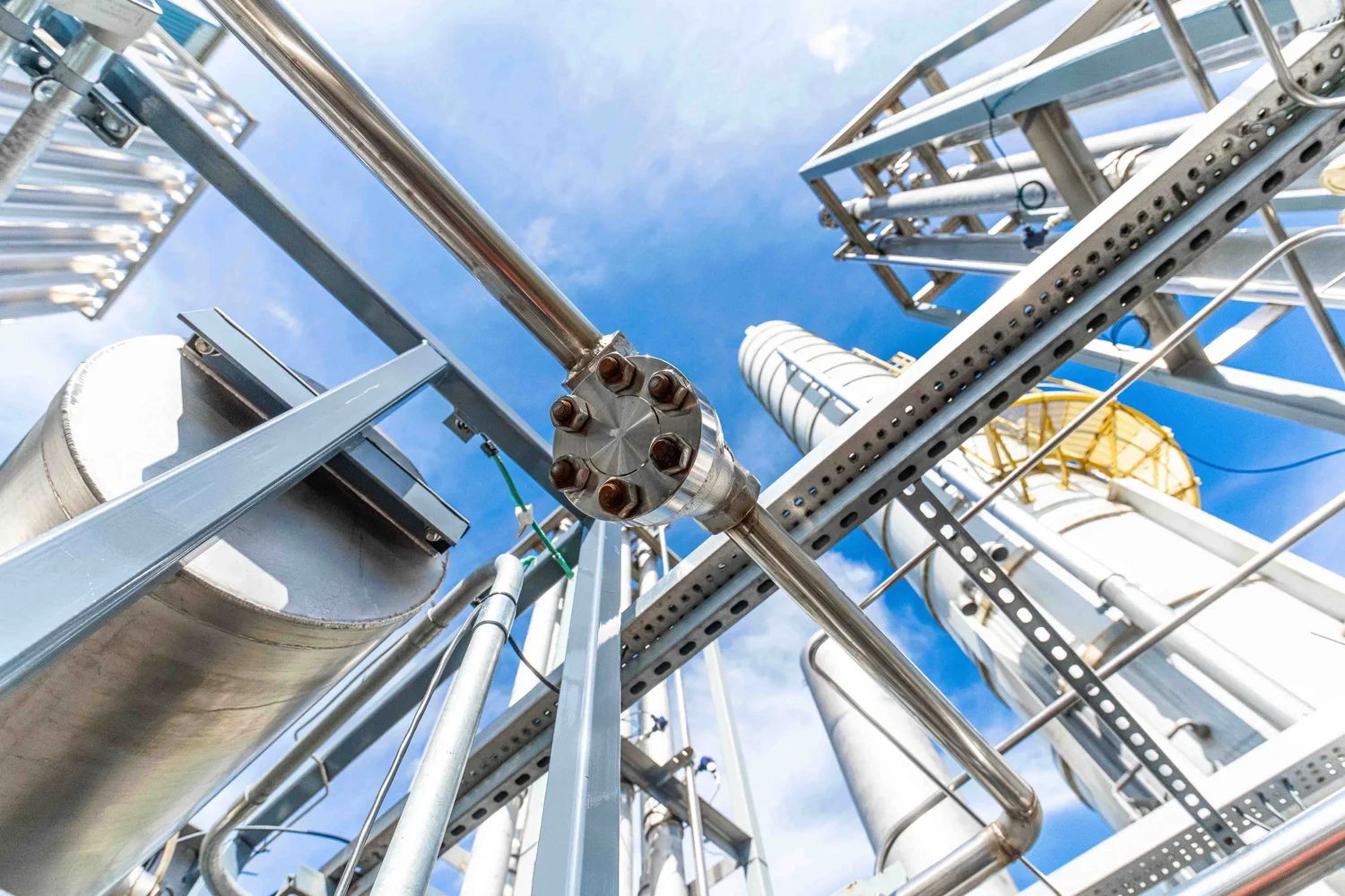Close-up industrial equipment photography of a bolted stainless steel pipe flange surrounded by vaporizer columns, structural steel and process piping, emphasizing engineered detail.